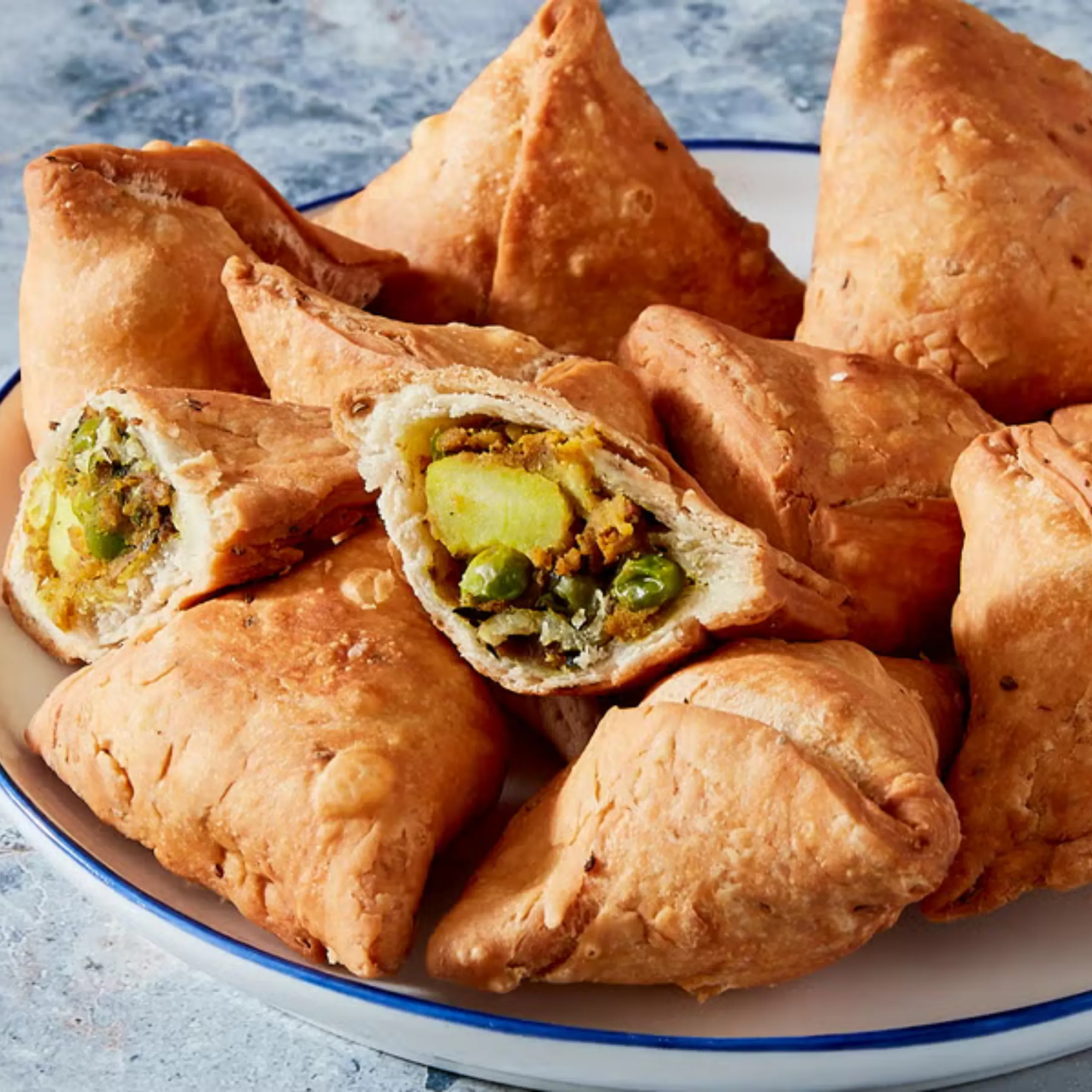 Golden-brown fried samosas on a white plate; one cut open to reveal mashed potato, peas and spices inside.