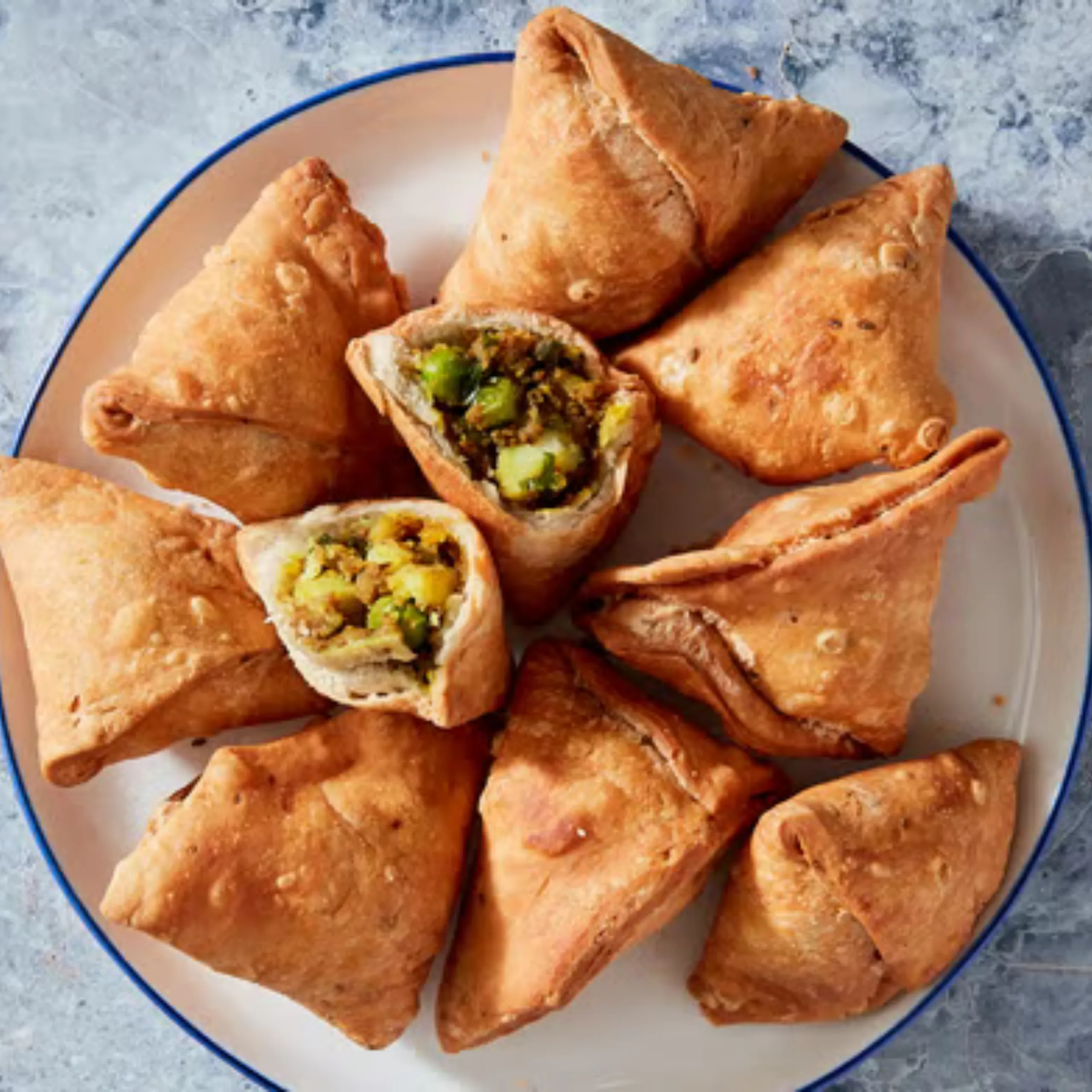 Golden fried samosas arranged on a white plate with blue rim, one cut open to reveal seasoned potato filling with peas and herbs