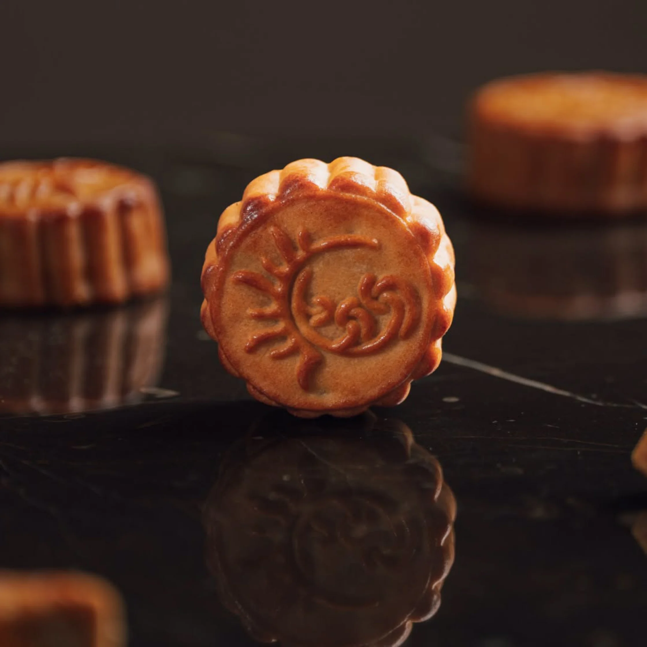 Golden-brown mooncake with an ornate embossed floral design stands upright on a reflective black surface, warmly lit against blurred pastries.