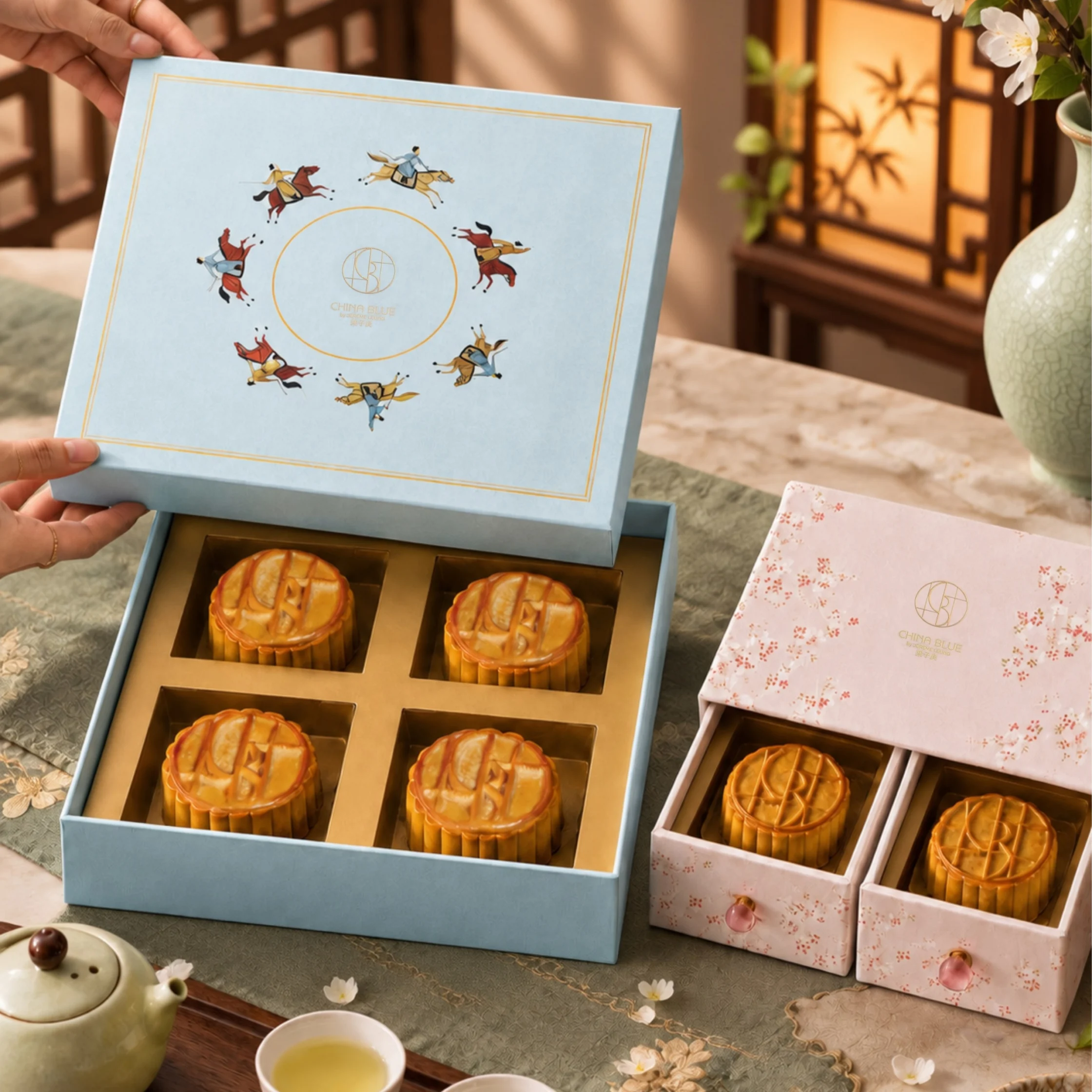 Open blue gift box displays four golden mooncakes beneath a crane design; pink floral boxes, tea, and warm sunlight create an elegant festive mood.