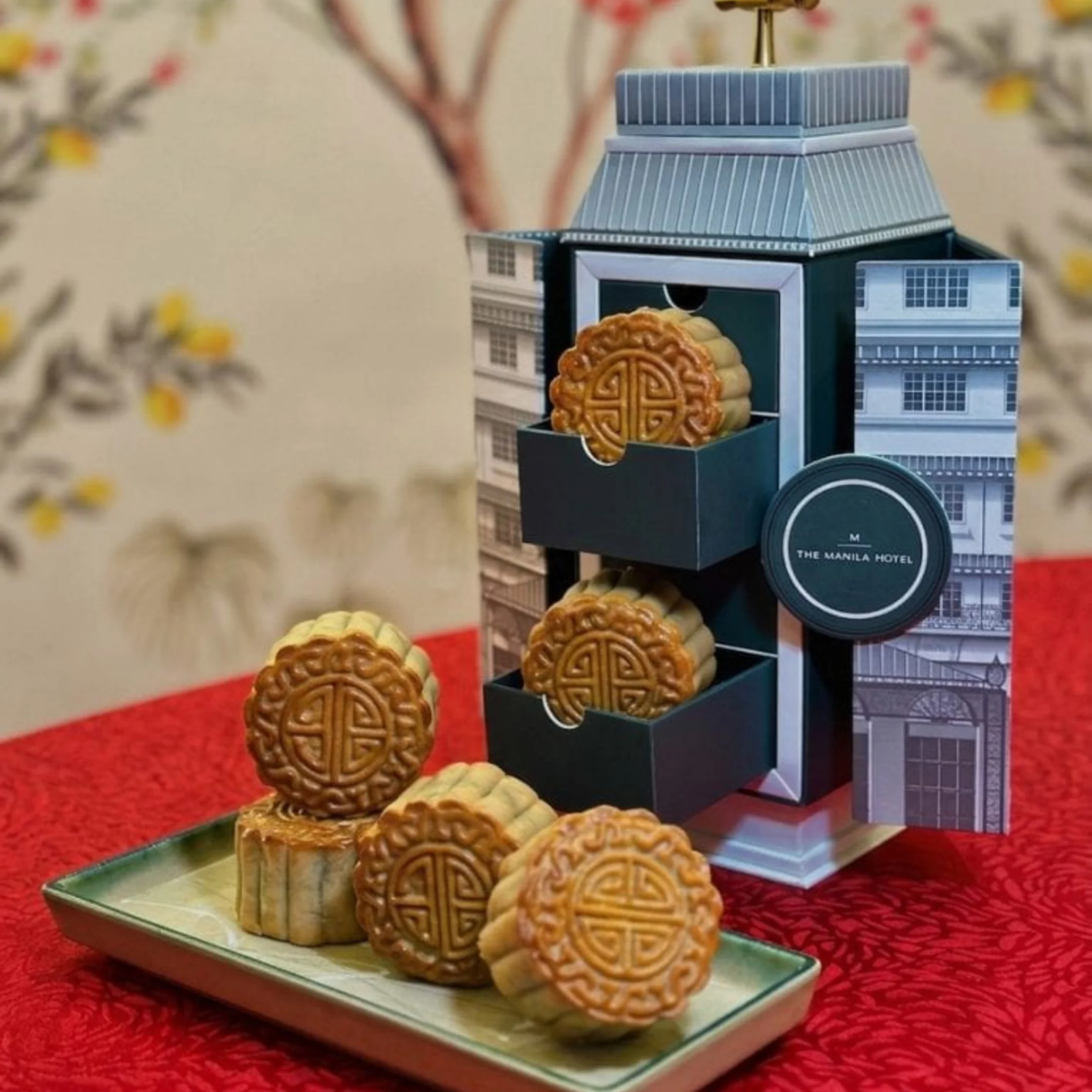 Decorative mooncake display shaped like a traditional hotel, with embossed pastries on a tray against red fabric and floral decor; festive, elegant mood.