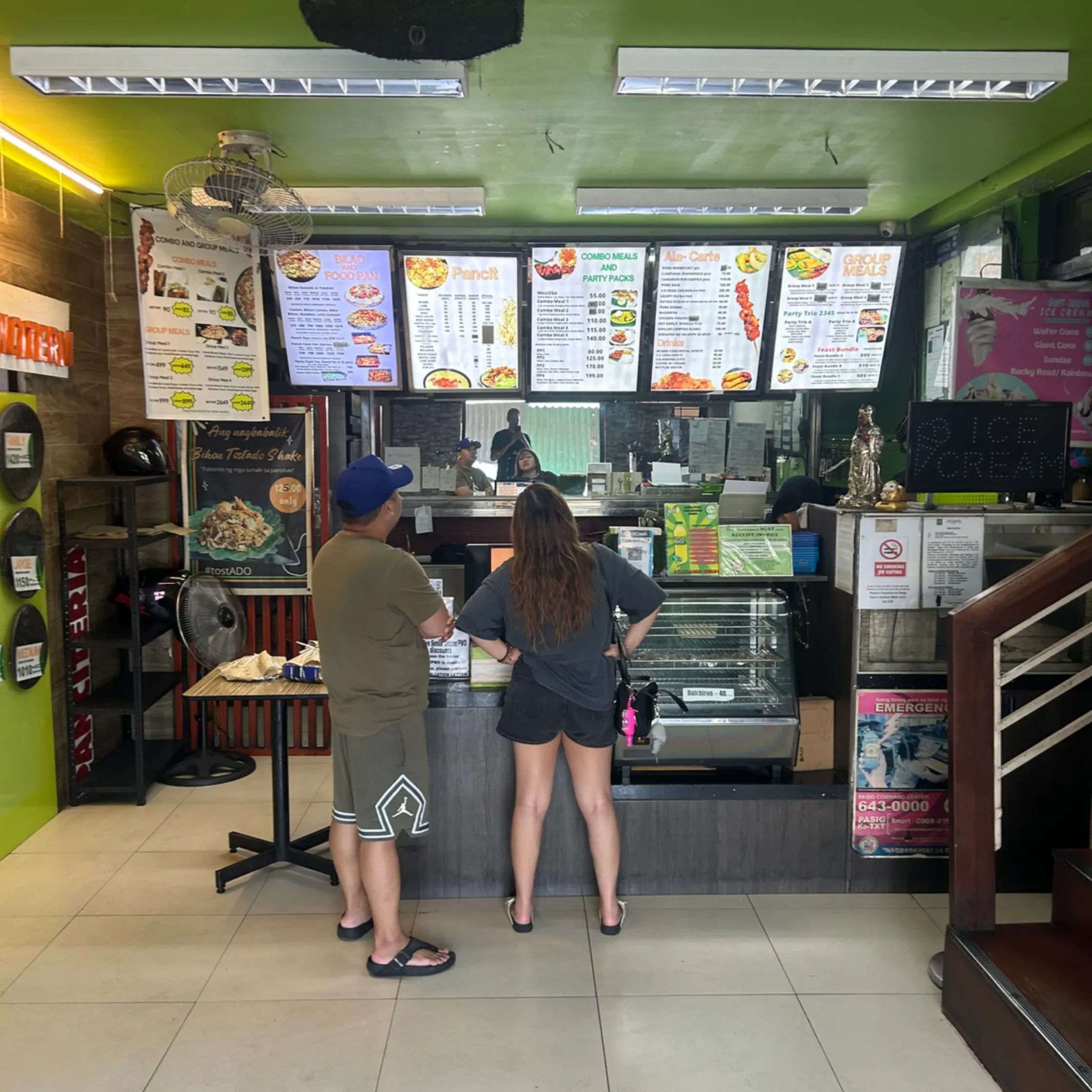 Two customers stand at a fast-food counter, facing the menu boards above the service area in a bright, green-walled shop.