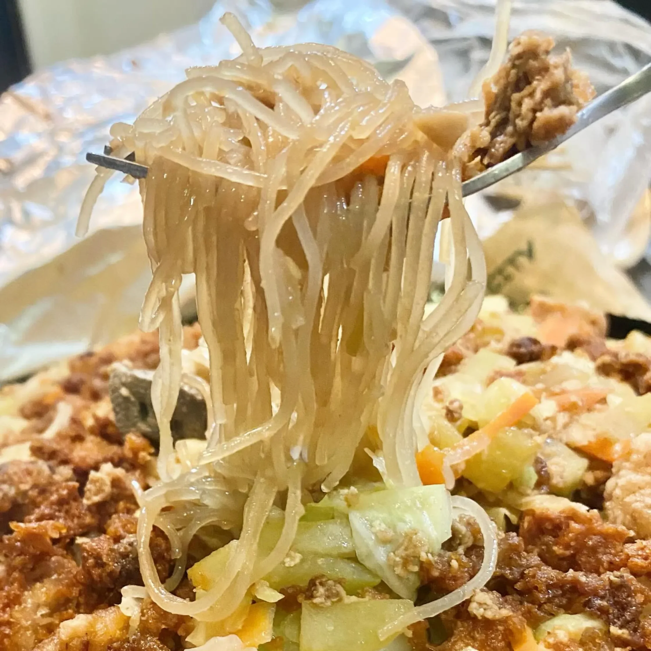 Close-up of a forkful of translucent glass noodles tangled with meat and vegetables in a saucy dish, highlighting a glossy texture and pale colors.