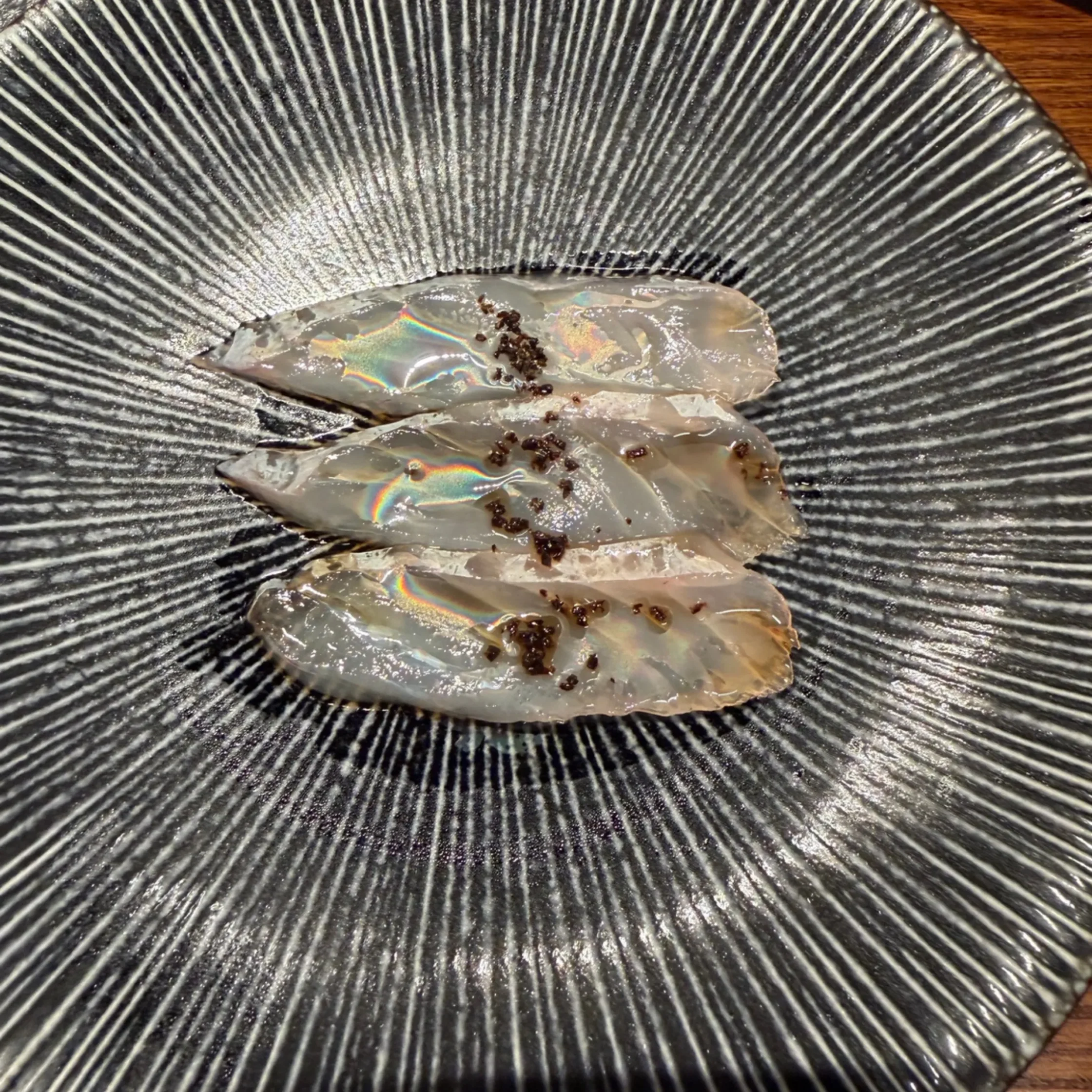Thin, iridescent slices of raw shellfish arranged on a dark, striped plate with a glossy finish.