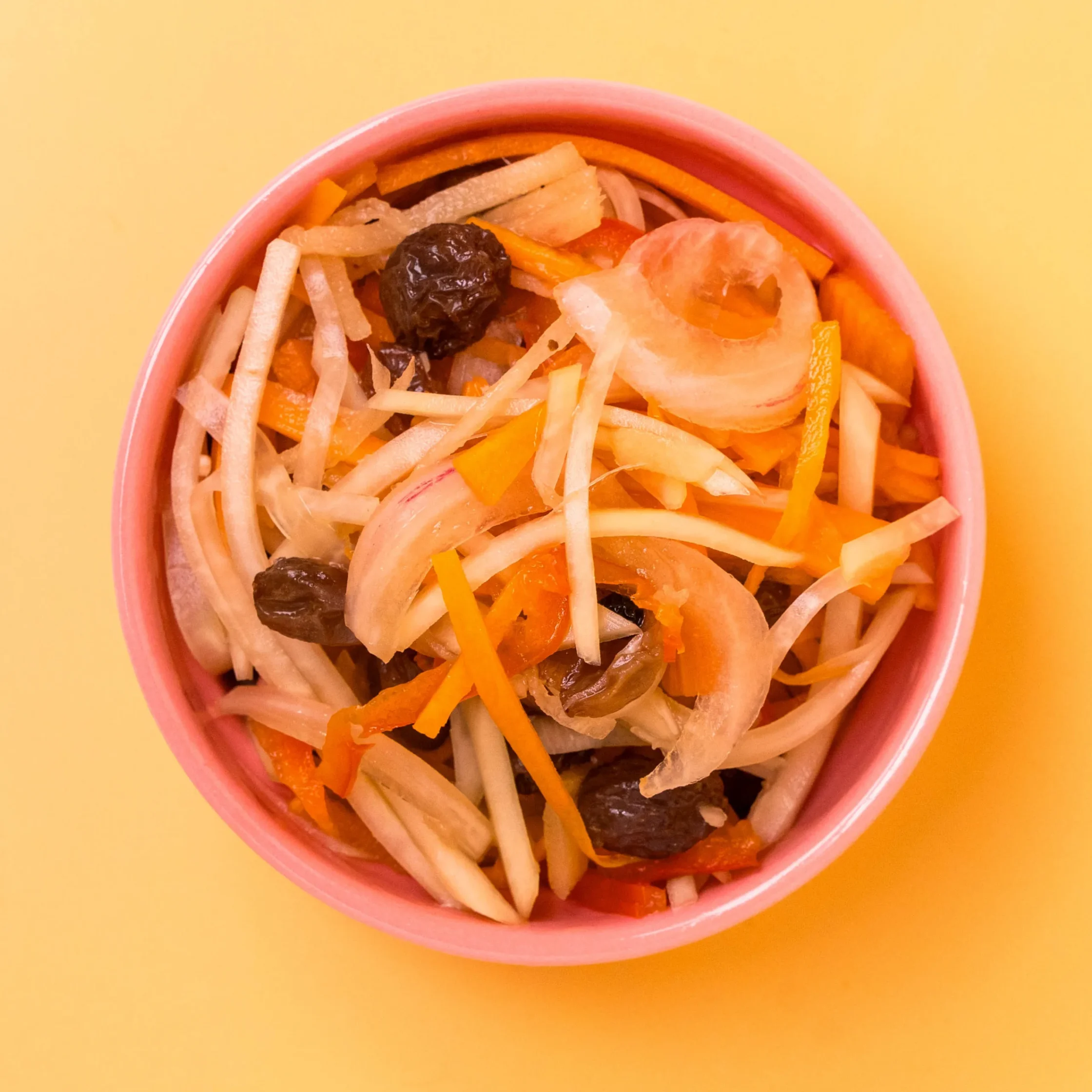 Pink bowl filled with shredded carrots, radish, and dark raisins on a yellow background, a colorful vegetable-fruit salad.
