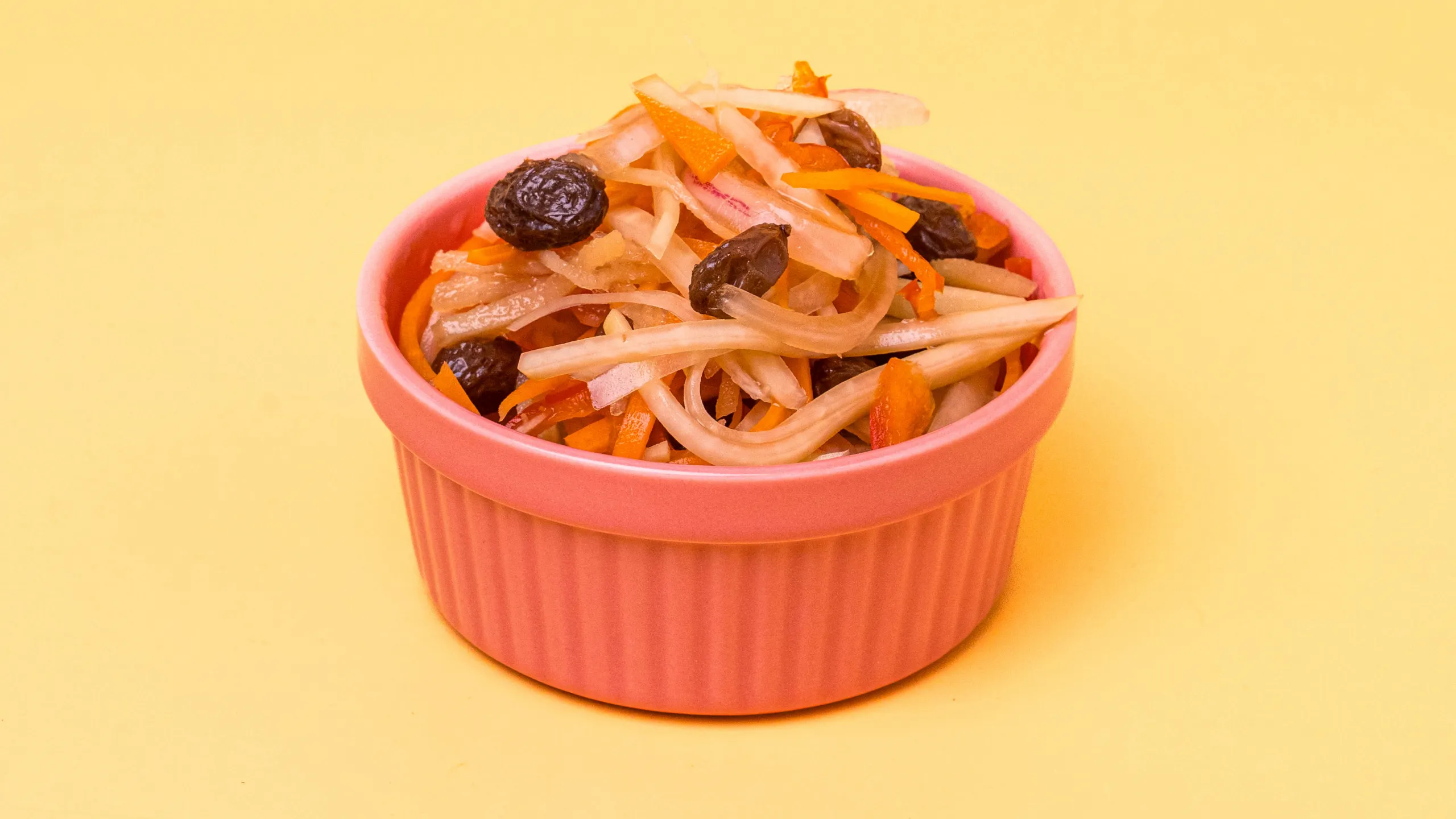 Pink ridged cup filled with a colorful shredded vegetable slaw (carrot, onion, potato-like root, prunes) against a yellow background