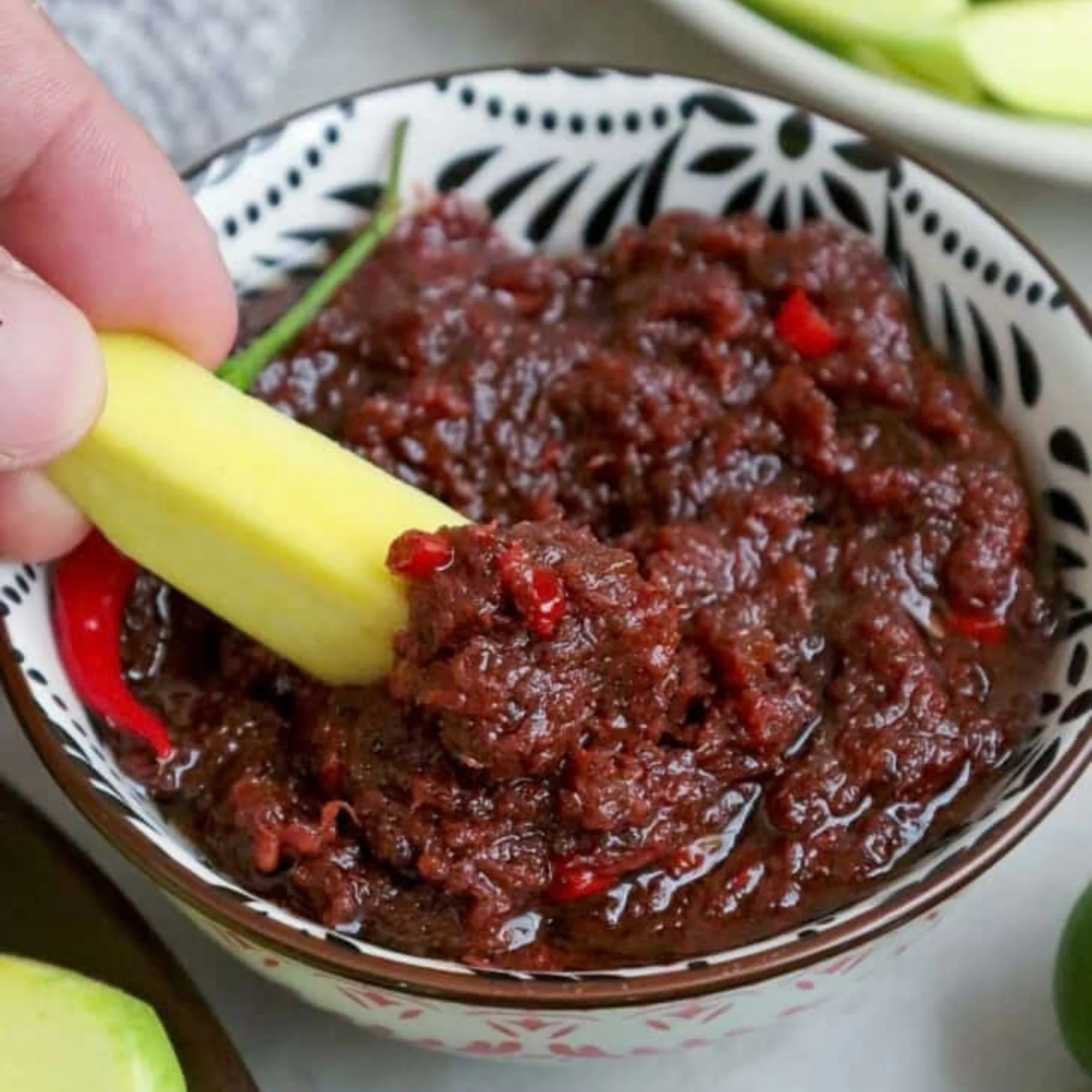 A hand dips a yellow vegetable stick into chunky dark-red salsa with visible pepper pieces, served in a patterned bowl; the scene feels fresh and appetizing.