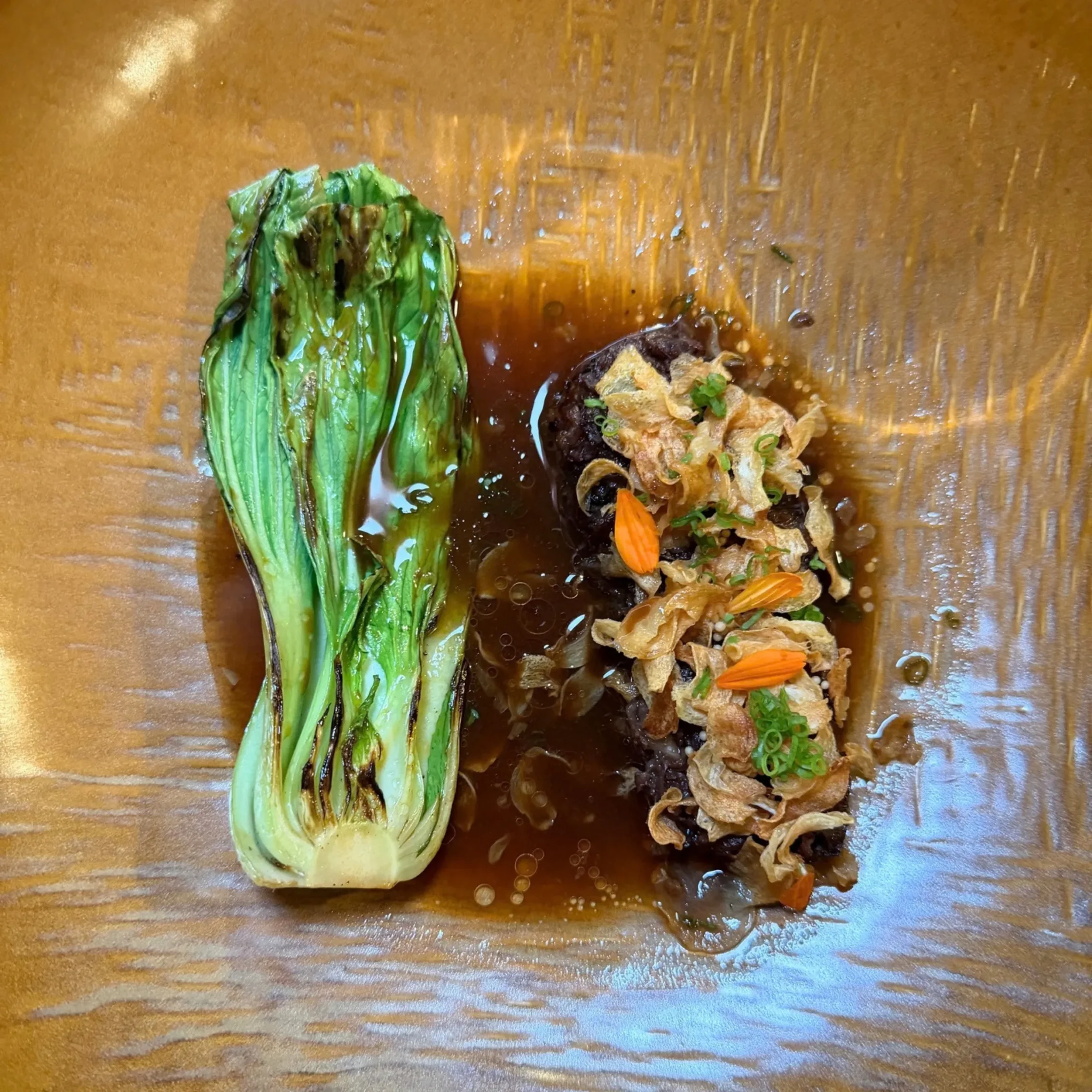 Overhead view of glossy grilled bok choy beside mushrooms, fried onions, almonds, and scallions in dark savory sauce on a wood plate.