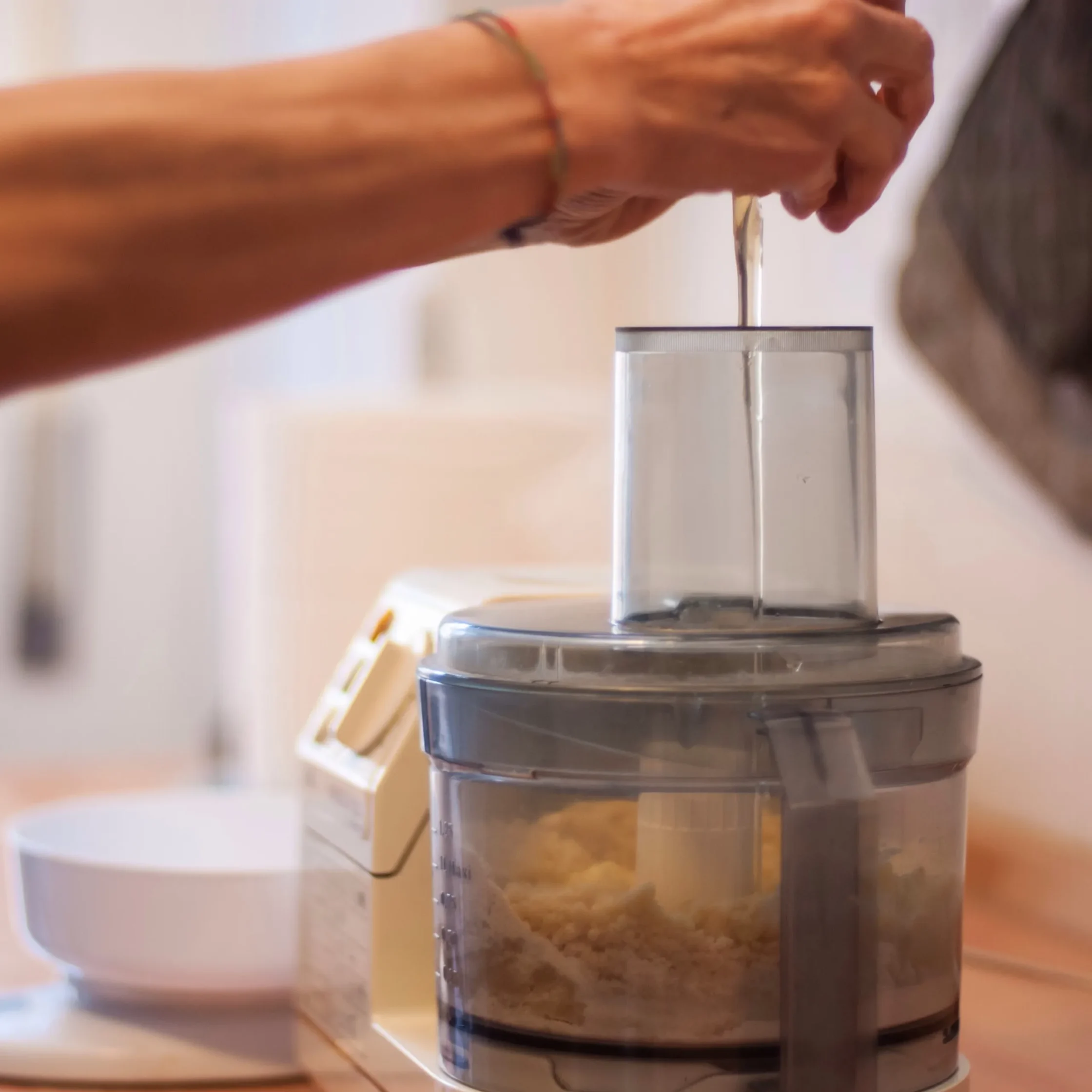 Person adds an ingredient through a food processor’s feed tube as dough mixes inside; a bowl and kitchen counter sit nearby