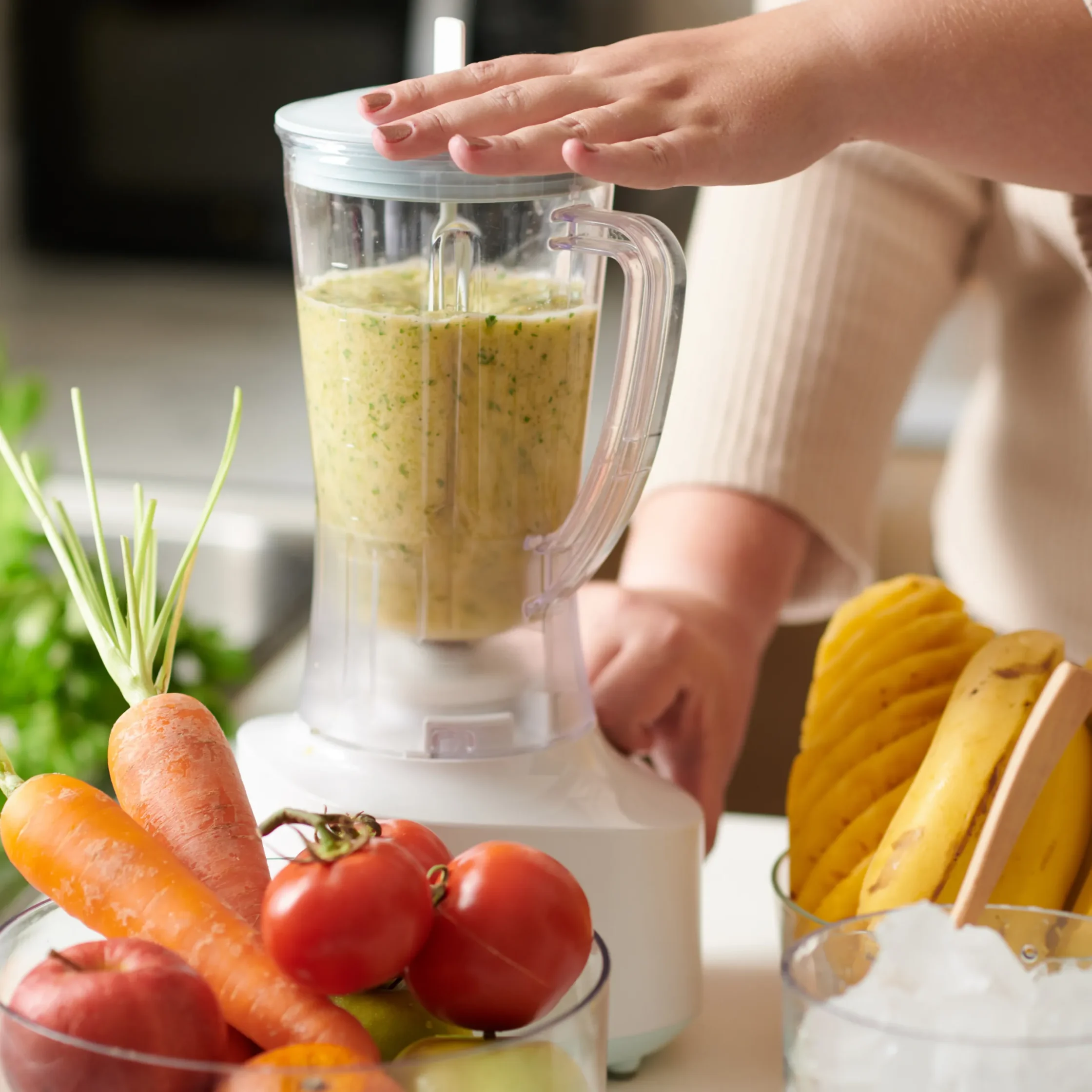 Person blends a green-flecked smoothie in a kitchen blender, surrounded by carrots, tomatoes, bananas, mango, and ice; fresh, healthy mood.