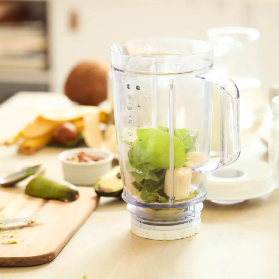 Blender jar filled with green apples, banana, leafy greens, and almonds sits on a kitchen counter beside chopped fruit, suggesting a fresh smoothie.