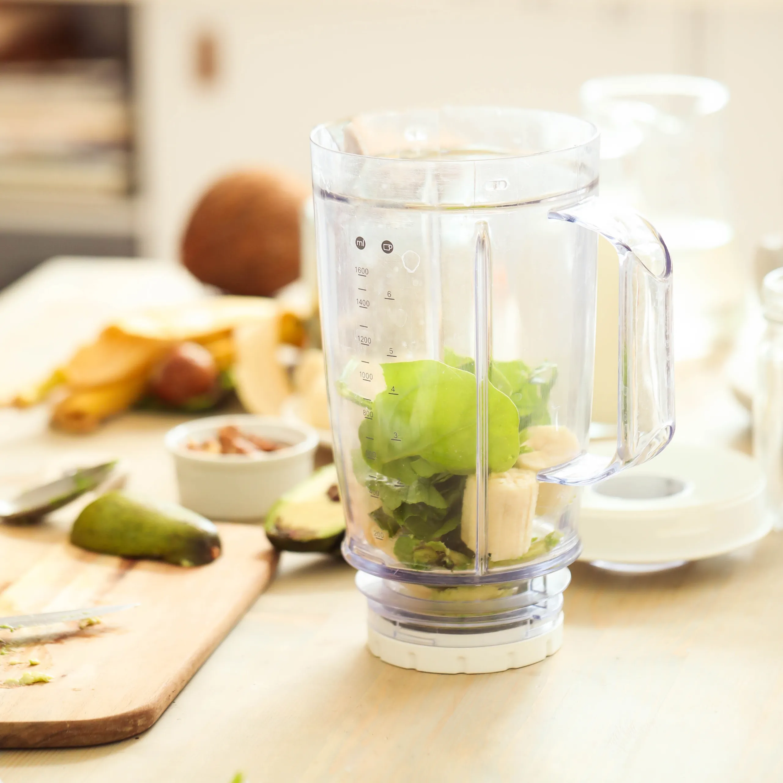 Blender jar filled with green apples, banana, leafy greens, and almonds sits on a kitchen counter beside chopped fruit, suggesting a fresh smoothie.