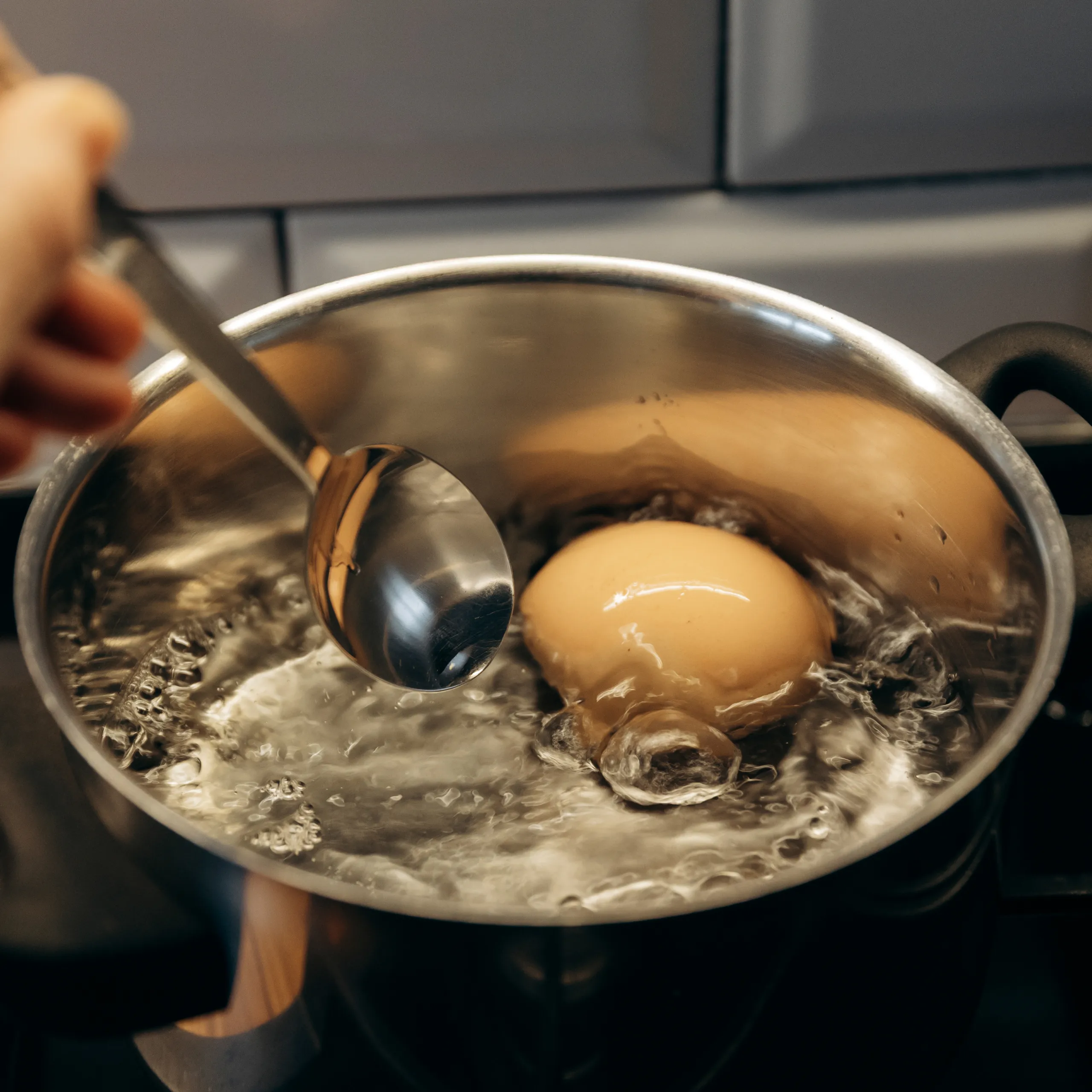 Eggs boiling in a stainless steel pot with a ladle and a hand nearby, steam rising from the water.
