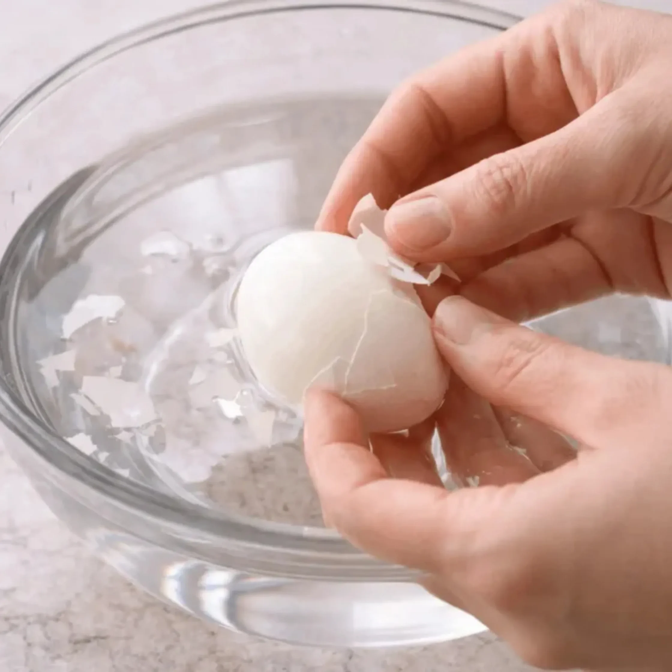 Close-up of hands breaking eggshells over a clear glass bowl on a light kitchen surface.