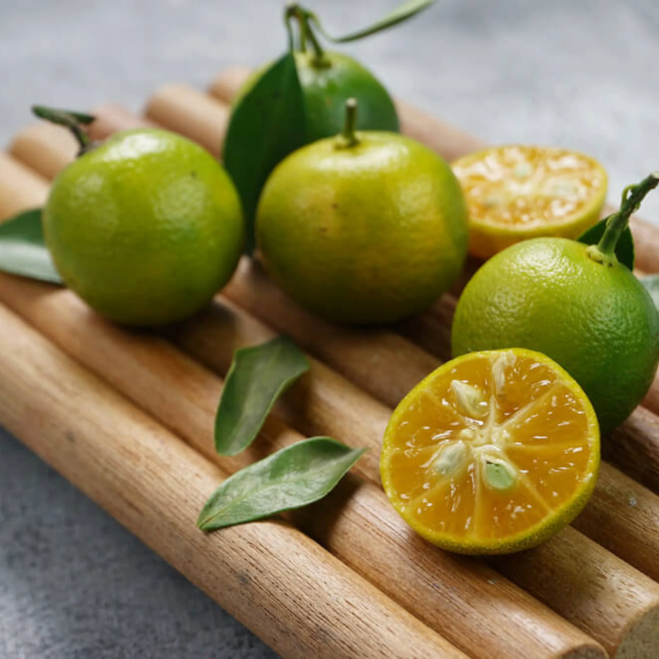 Green and yellow citrus fruits, whole and halved to reveal juicy, golden flesh, rest with fresh leaves on a wooden board, evoking a bright, natural feel.