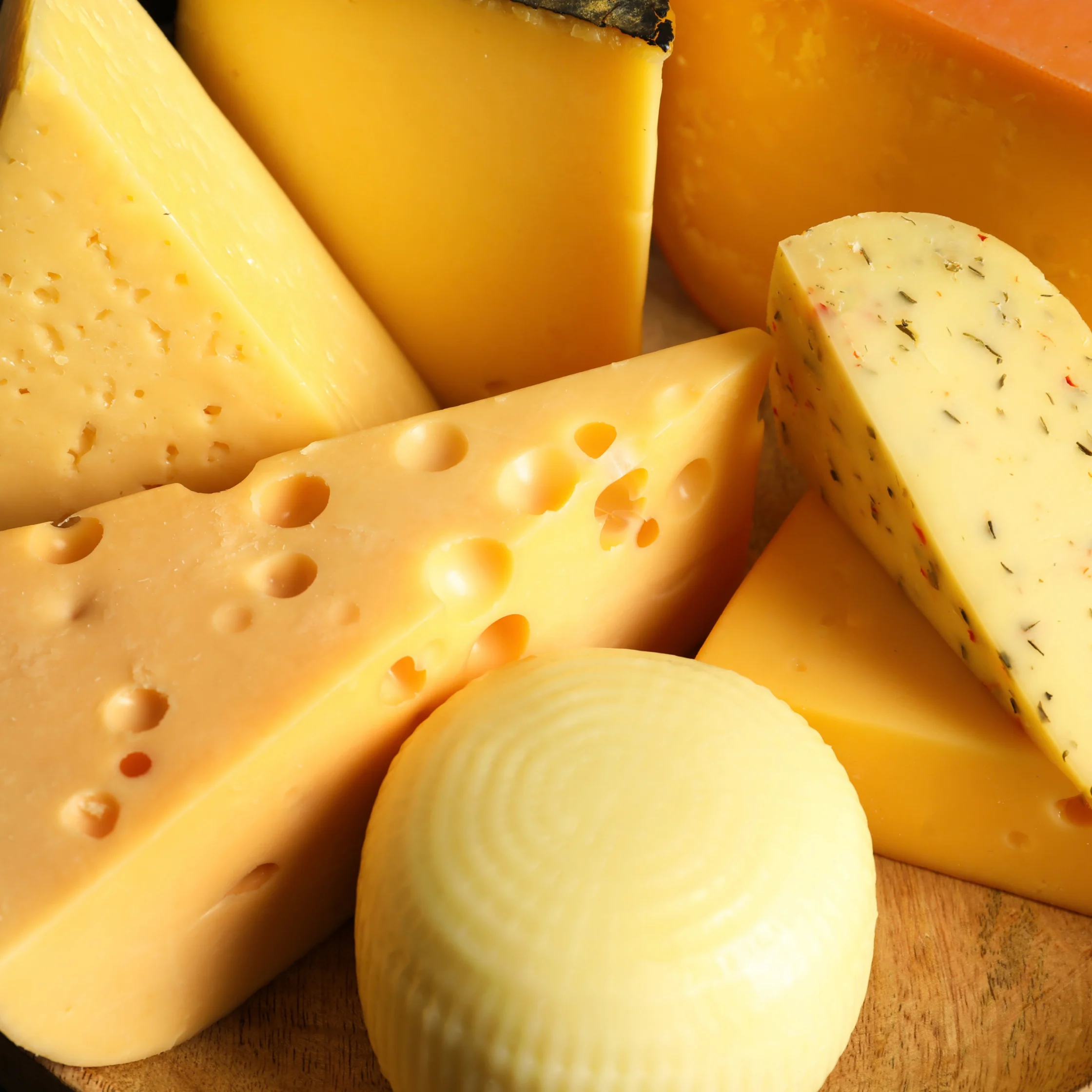 Close-up of assorted cheeses, including Swiss, cheddar, herb-speckled, and soft round varieties, arranged on a wooden board in warm light.