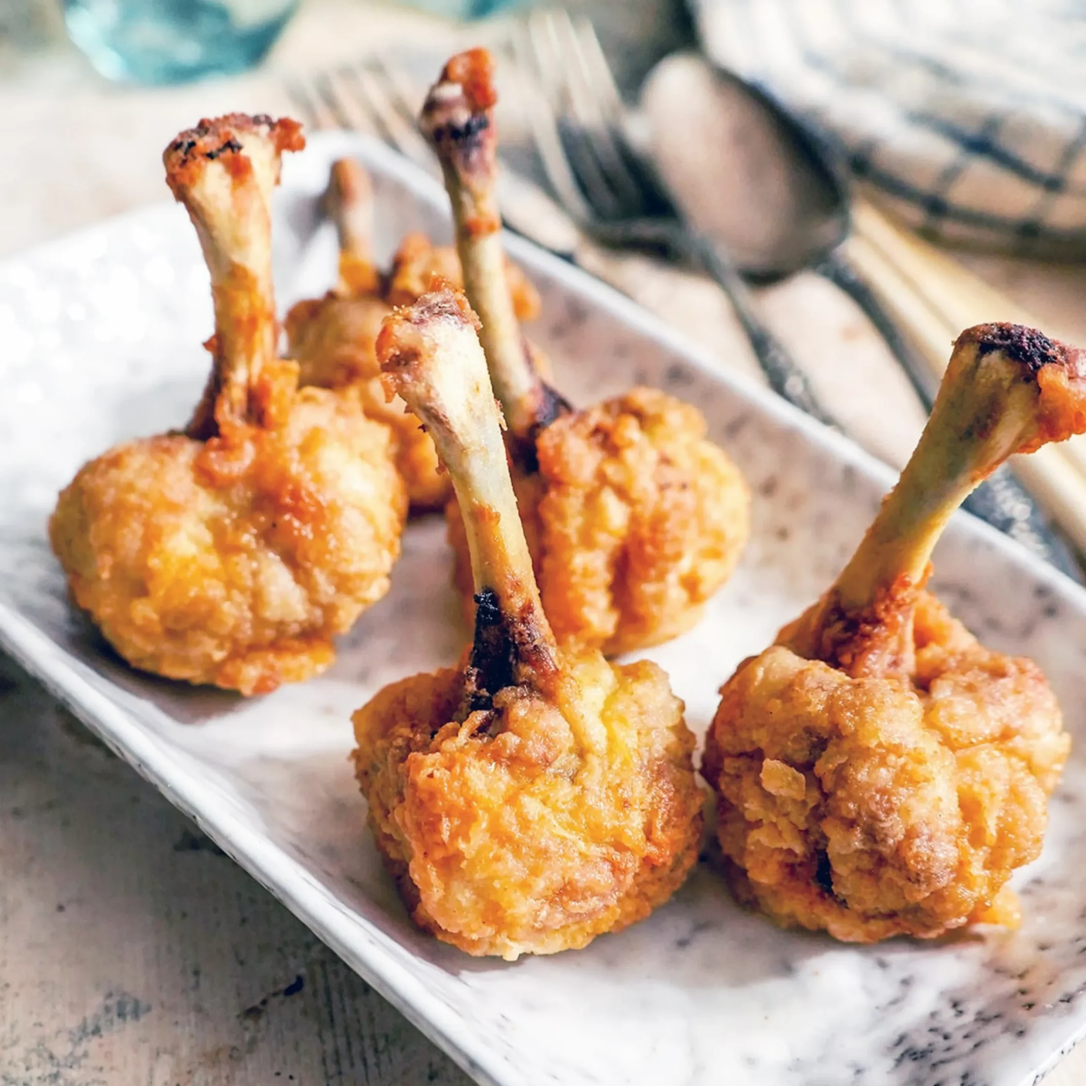 Golden-brown fried chicken lollipops arranged on a white platter, with water glasses and a napkin in the softly blurred background.