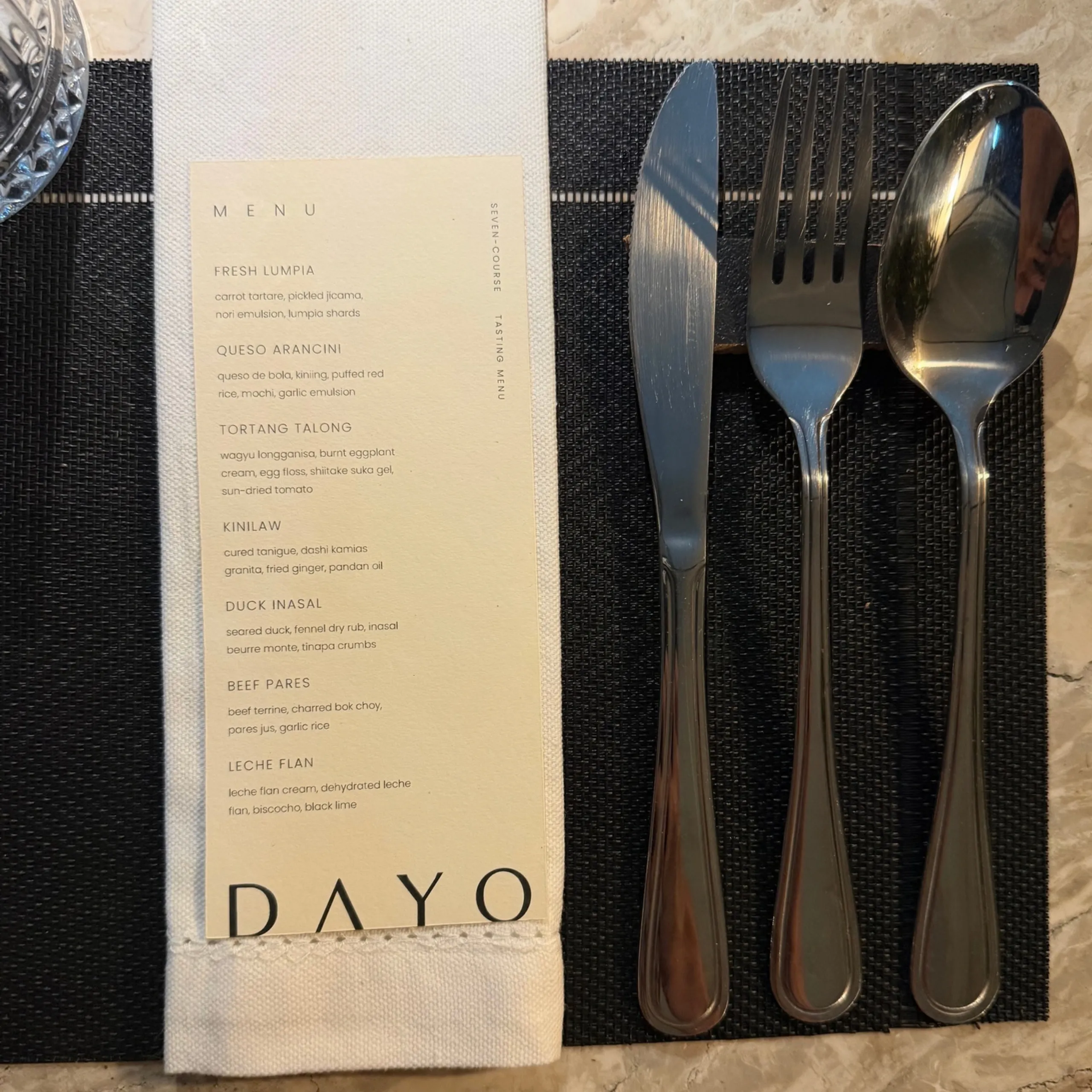 Elegant restaurant table setting with a DAYO menu, fork, knife, and spoon on a black placemat, conveying a refined, welcoming dining mood.