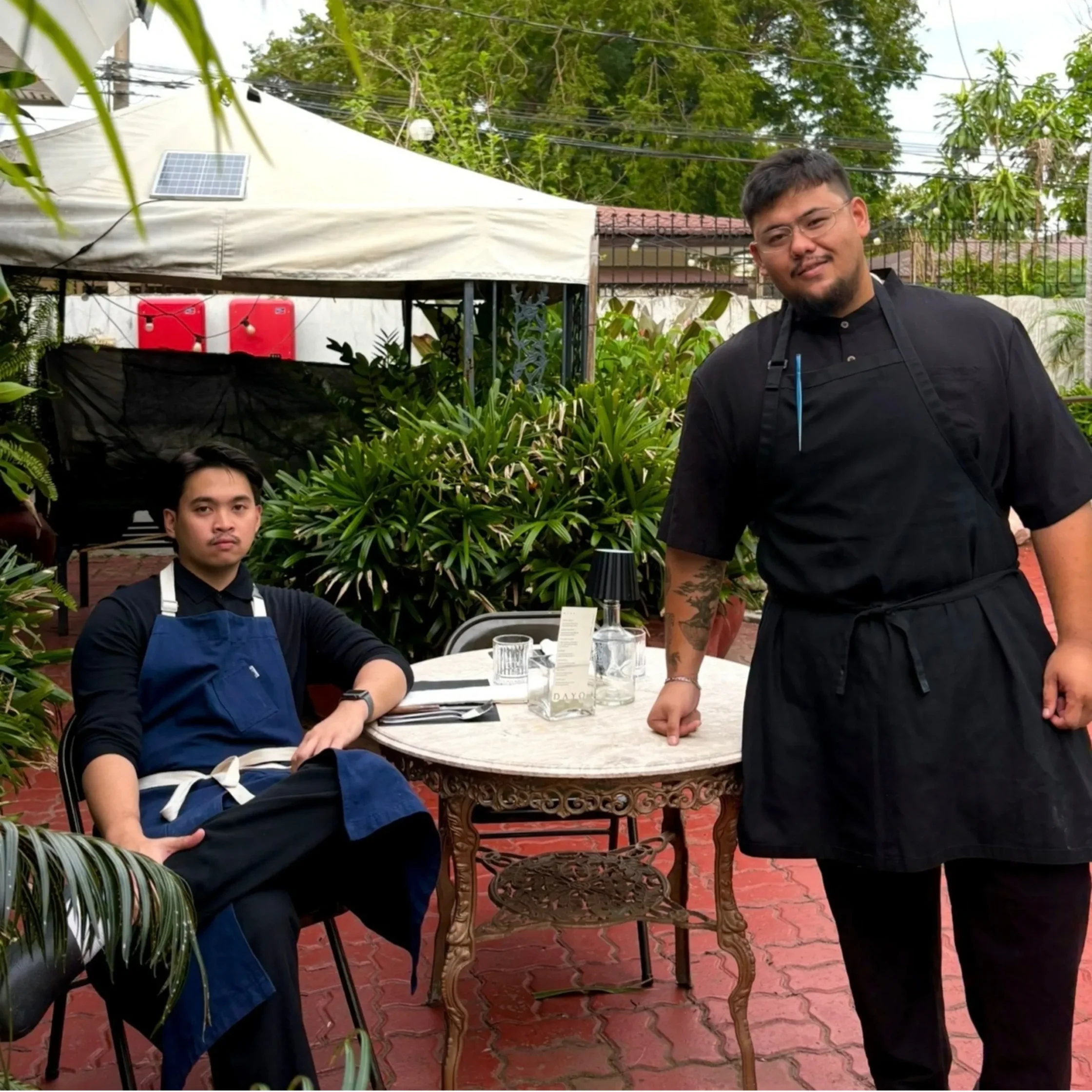 Two chefs in black uniforms pose on a lush outdoor restaurant patio, one seated and one standing beside a set table under a canopy.