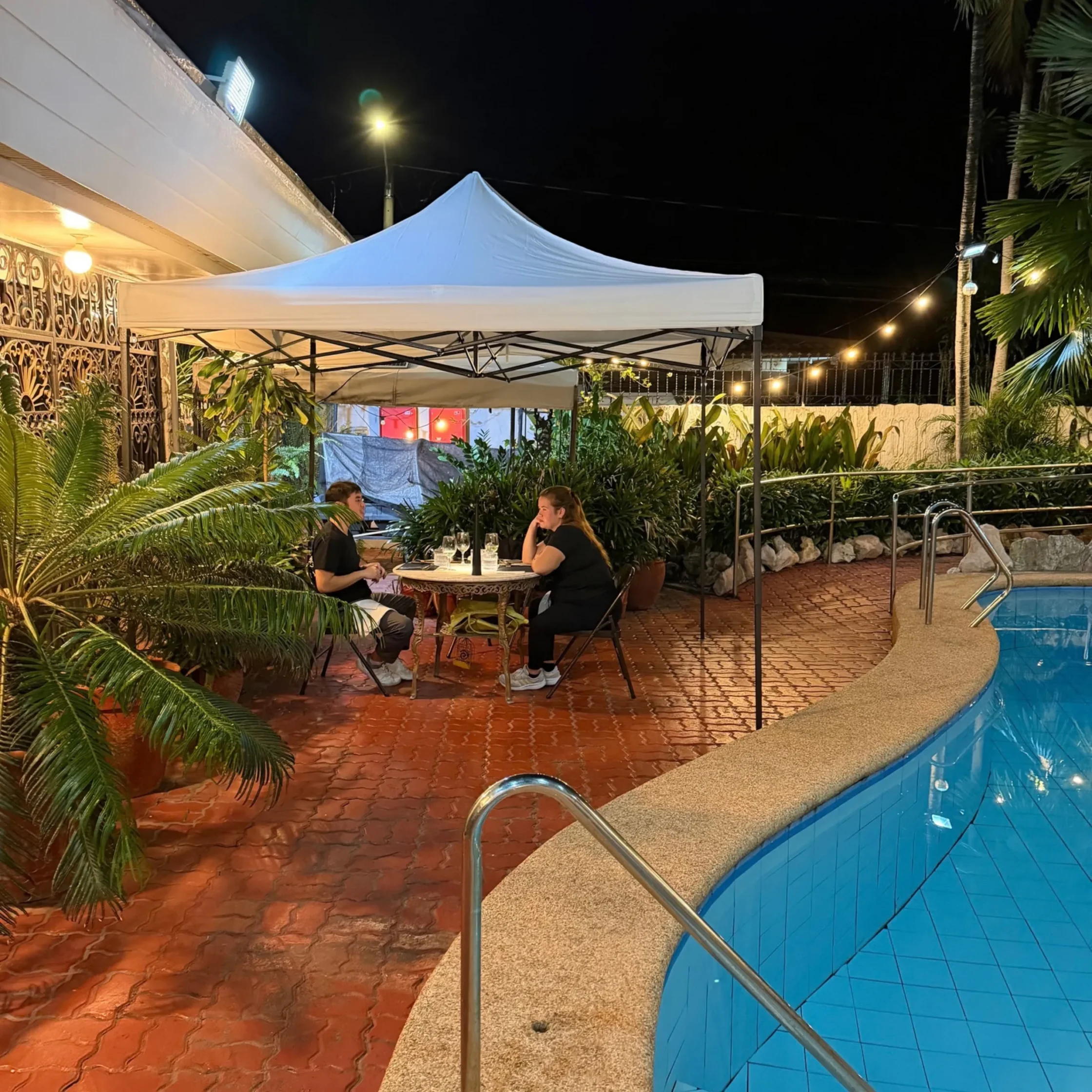 Nighttime patio with two people dining beneath a white canopy, surrounded by tropical plants, string lights, and a blue swimming pool.