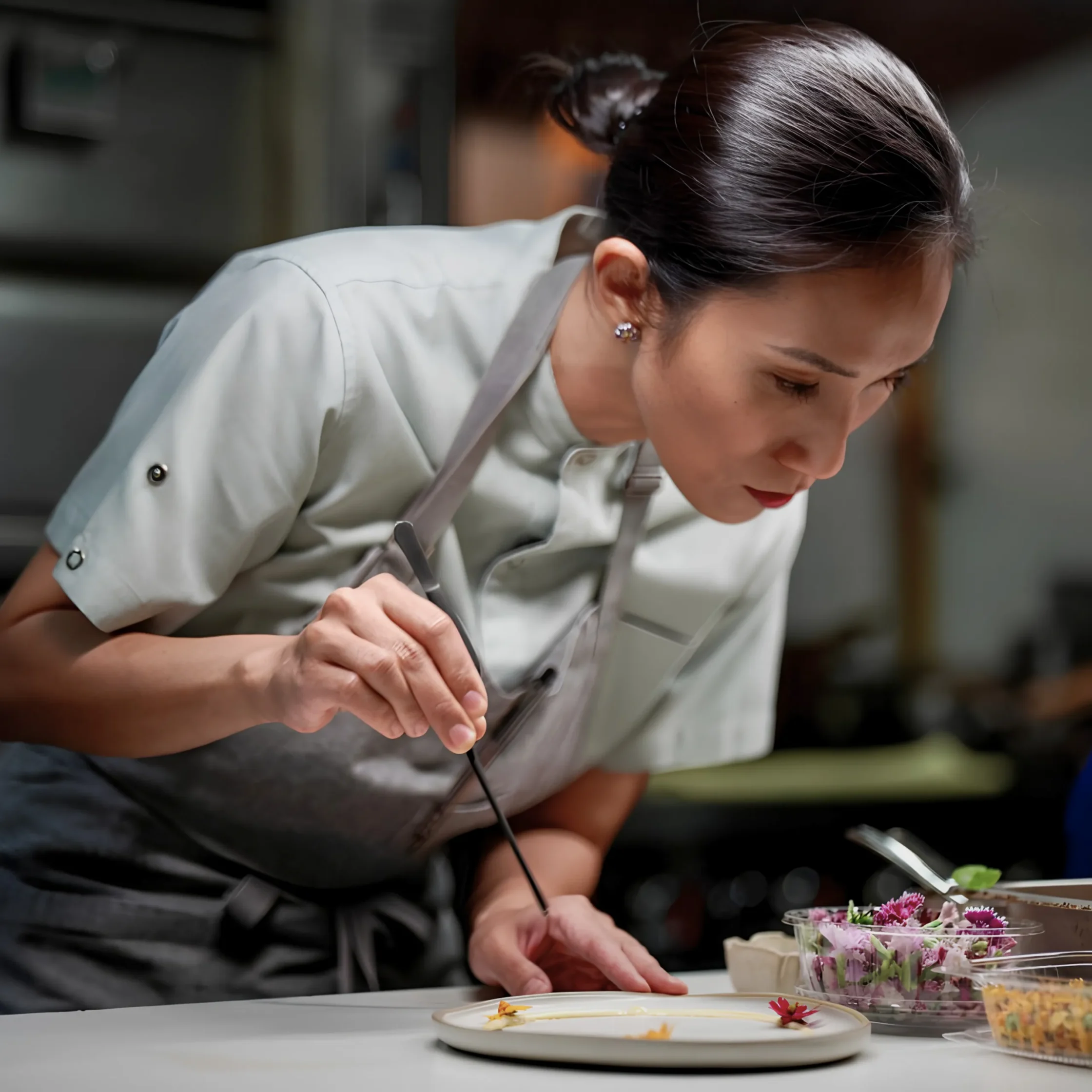 Person in a light apron concentrates on trimming flower petals at a kitchen counter with trays of flowers nearby.