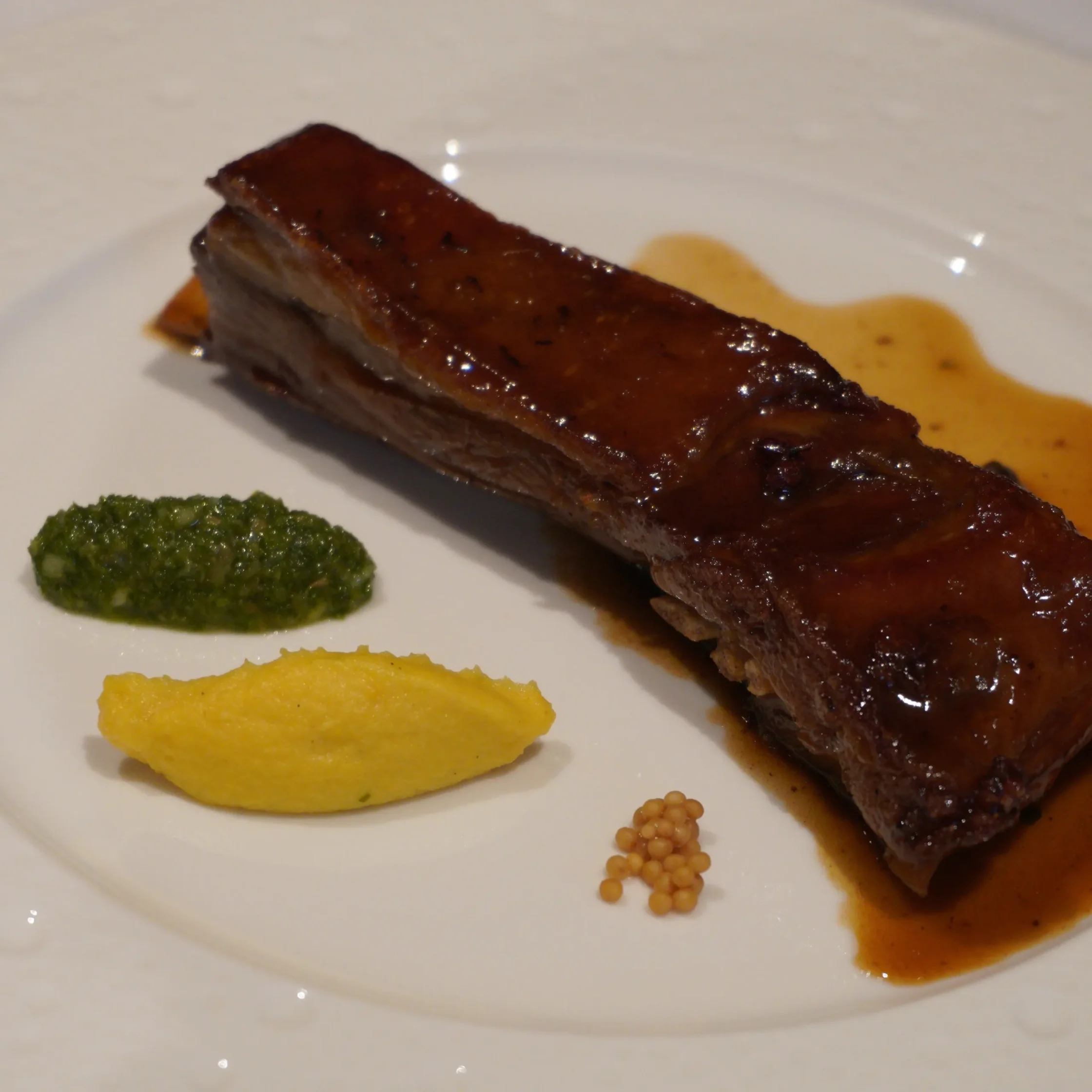 Slice of glazed meat on a white plate with green herb paste, yellow puree, and small mustard seeds nearby.