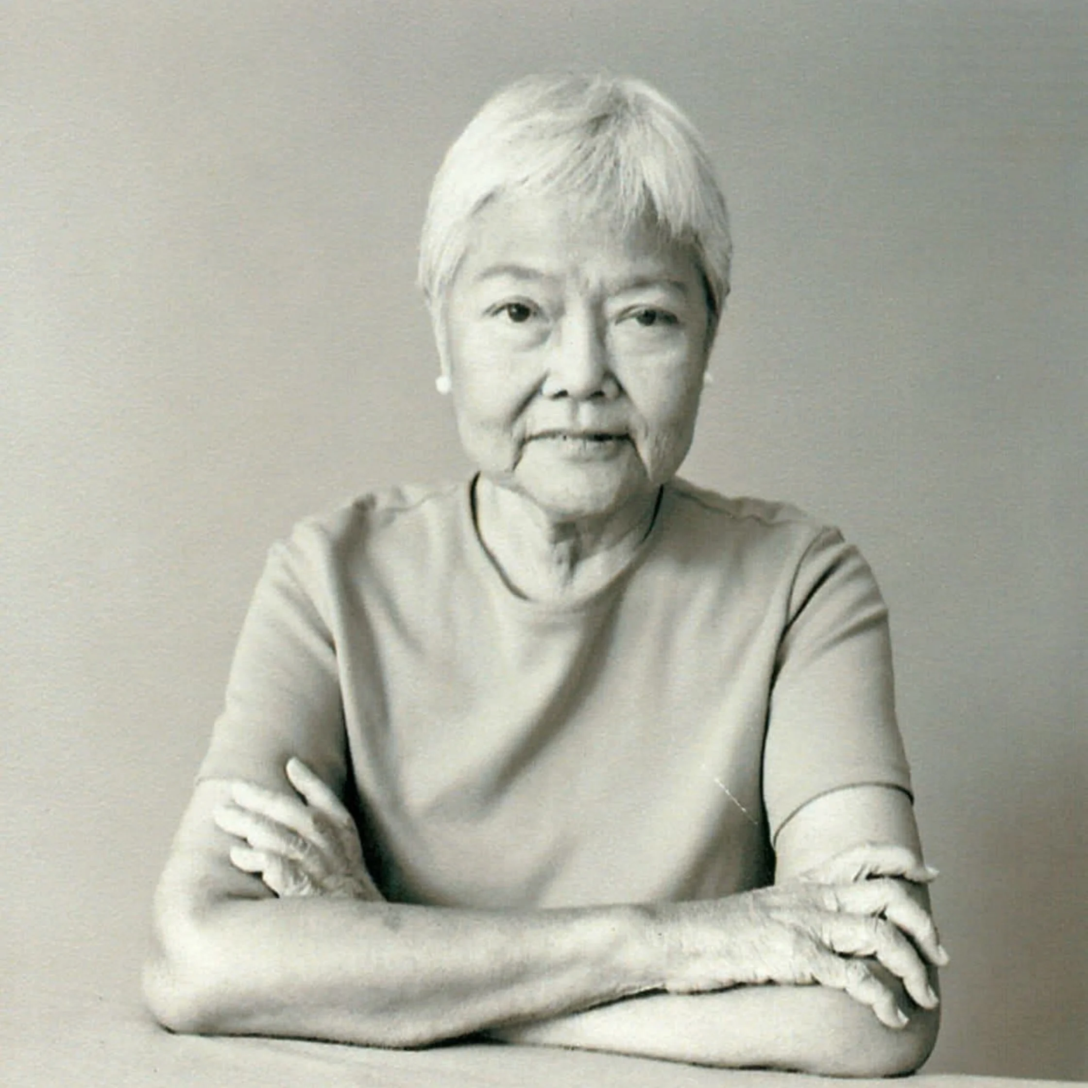 Black-and-white portrait of an older woman with short white hair, seated with arms folded, looking calmly at the camera against a plain backdrop.