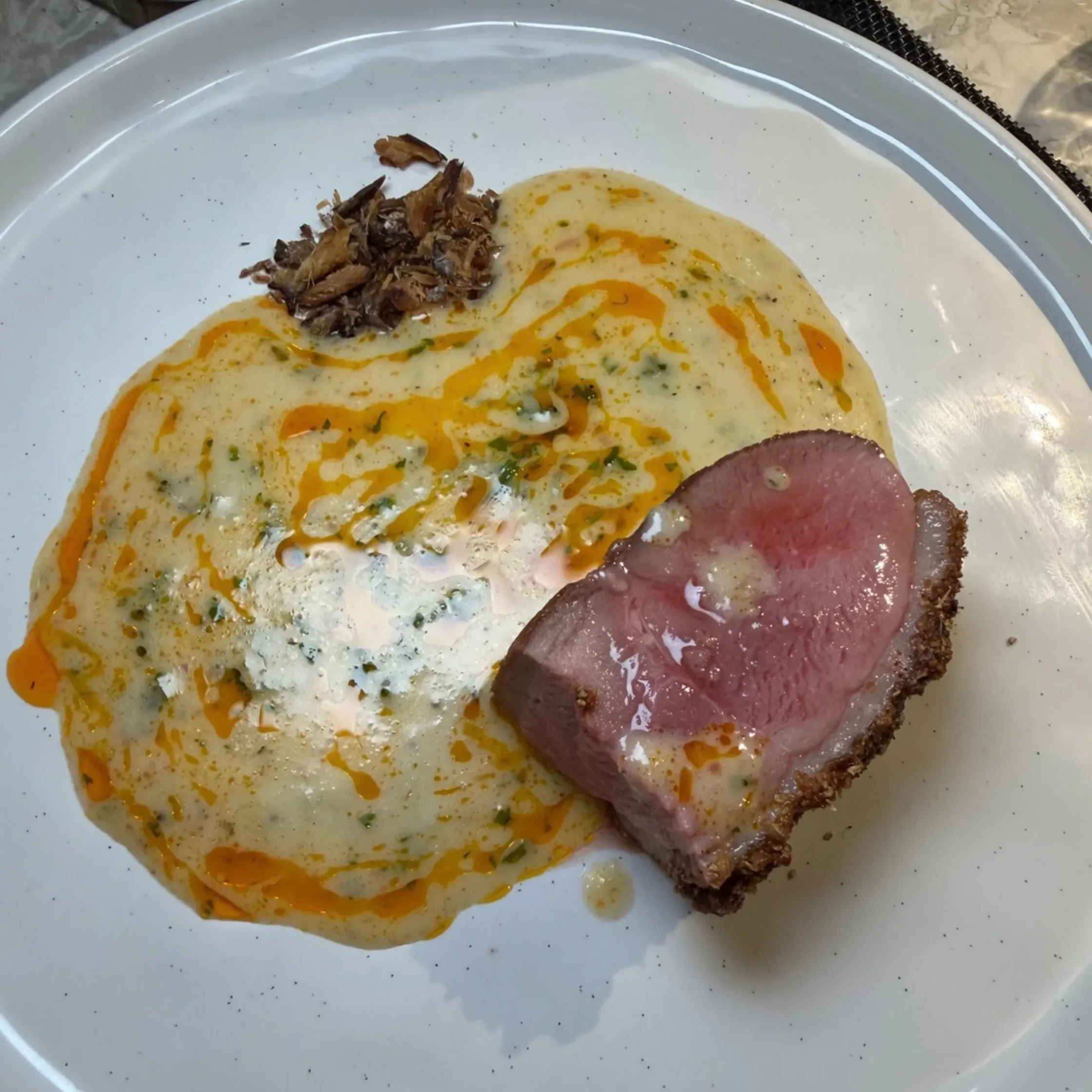 A gourmet plate features sliced medium-rare beef with crisp seared edges, creamy herb sauce, orange oil drizzle, and a crunchy garnish.