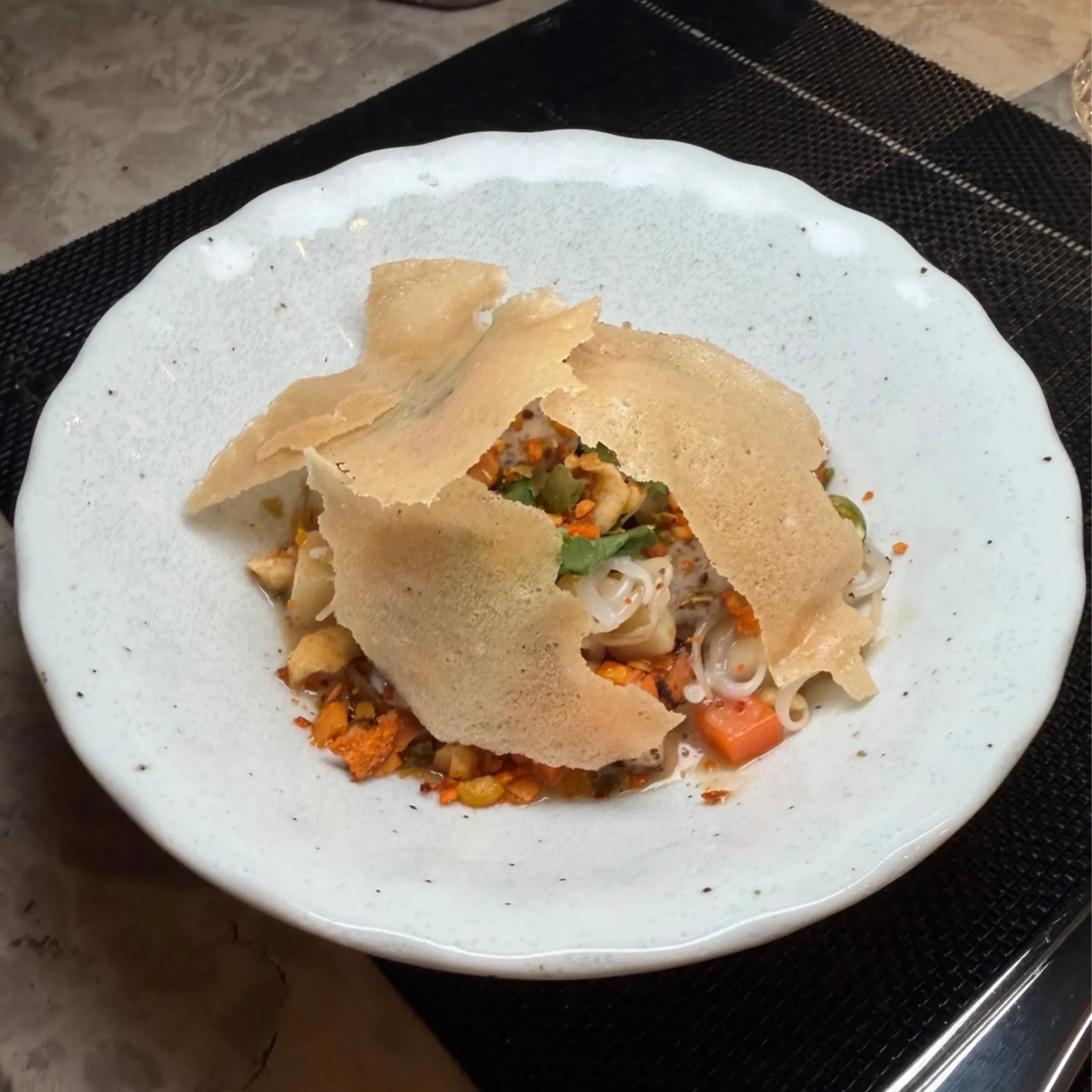 A white ceramic plate holds noodles, vegetables, and savory toppings beneath crisp, broken flatbread; a colorful, appetizing meal on a dark placemat.
