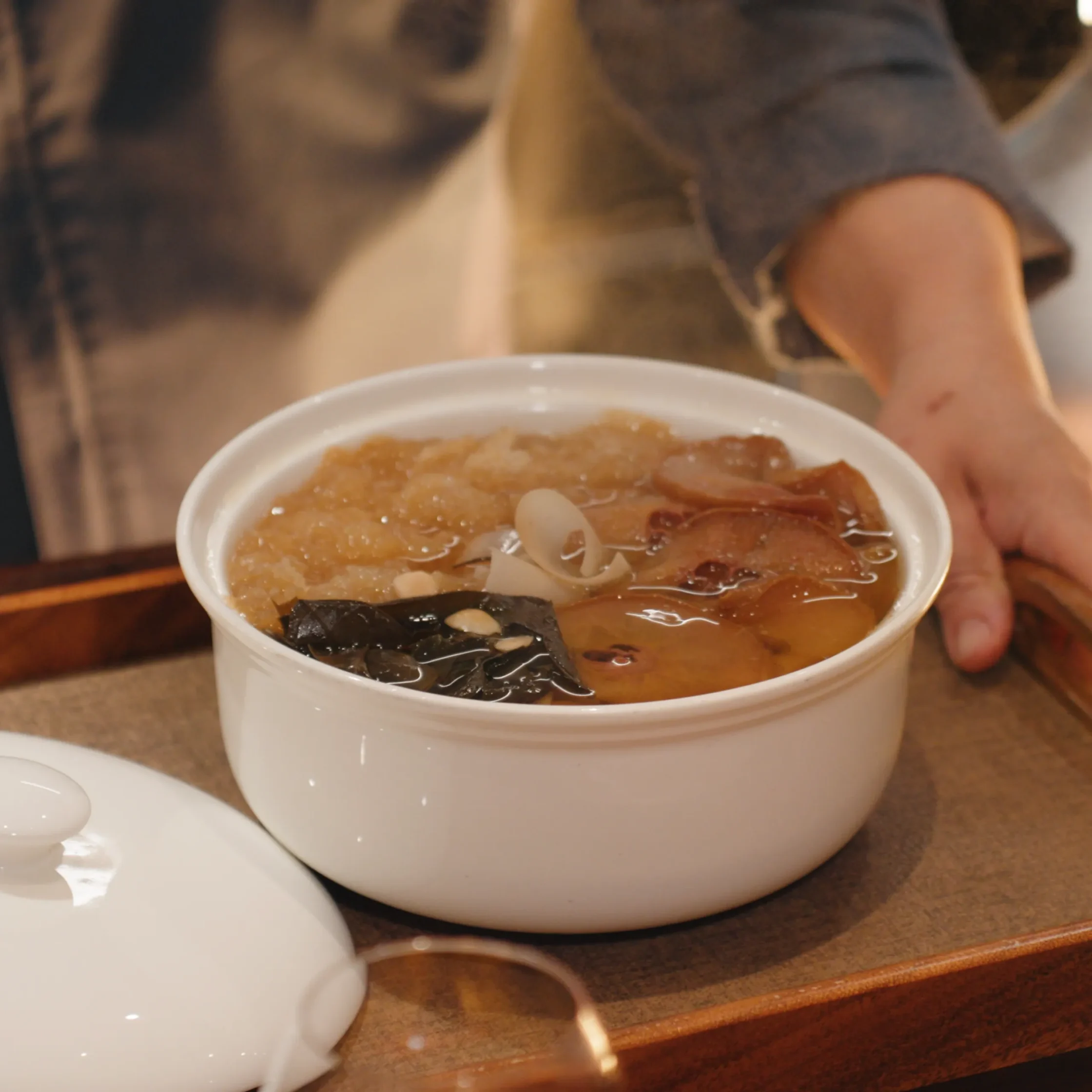 Cup of noodle soup with seaweed and noodles on a wooden table; a person’s hand is near the cup.