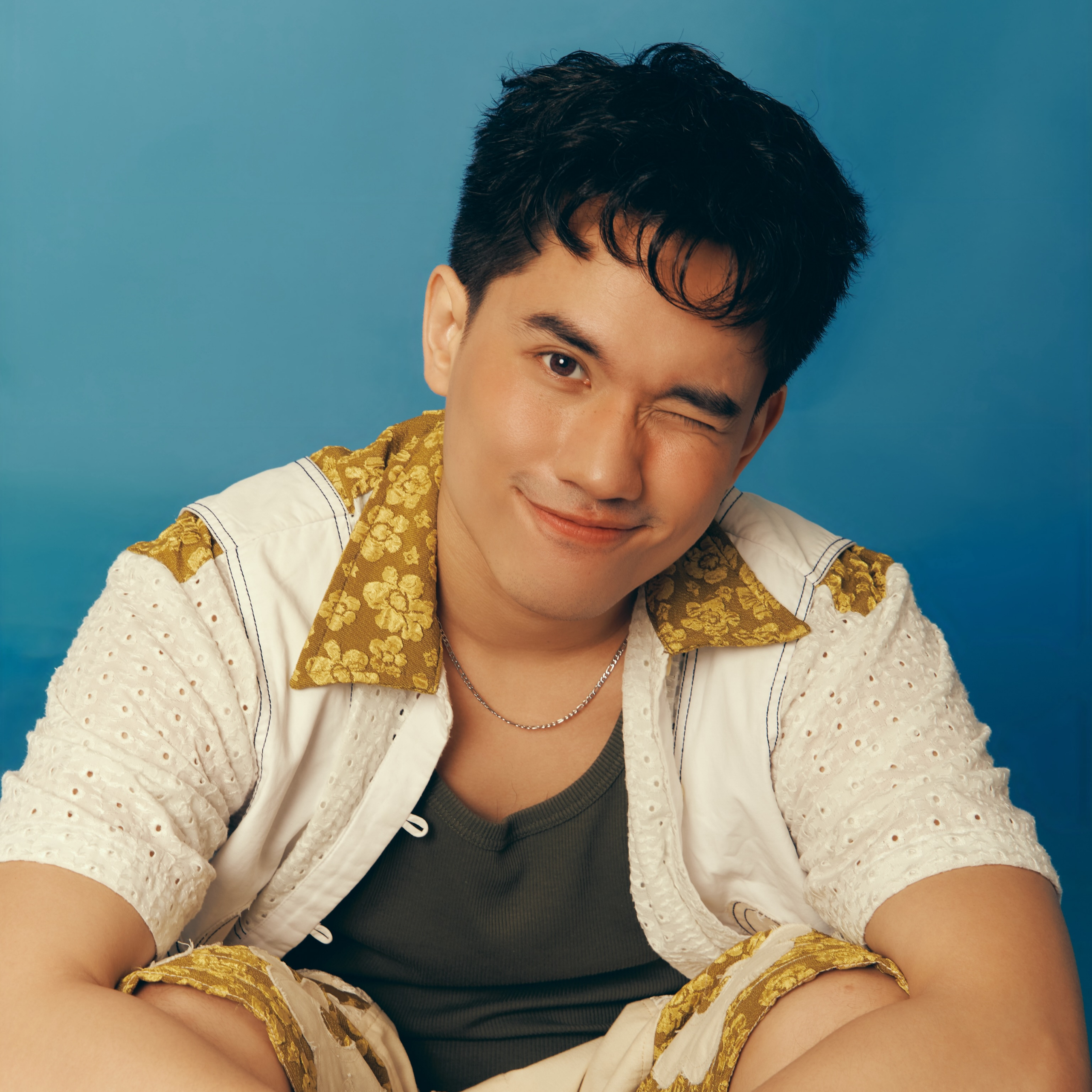 Portrait of a young man winking and smiling at the camera, wearing a cream patterned shirt over a dark tank top against a blue background.