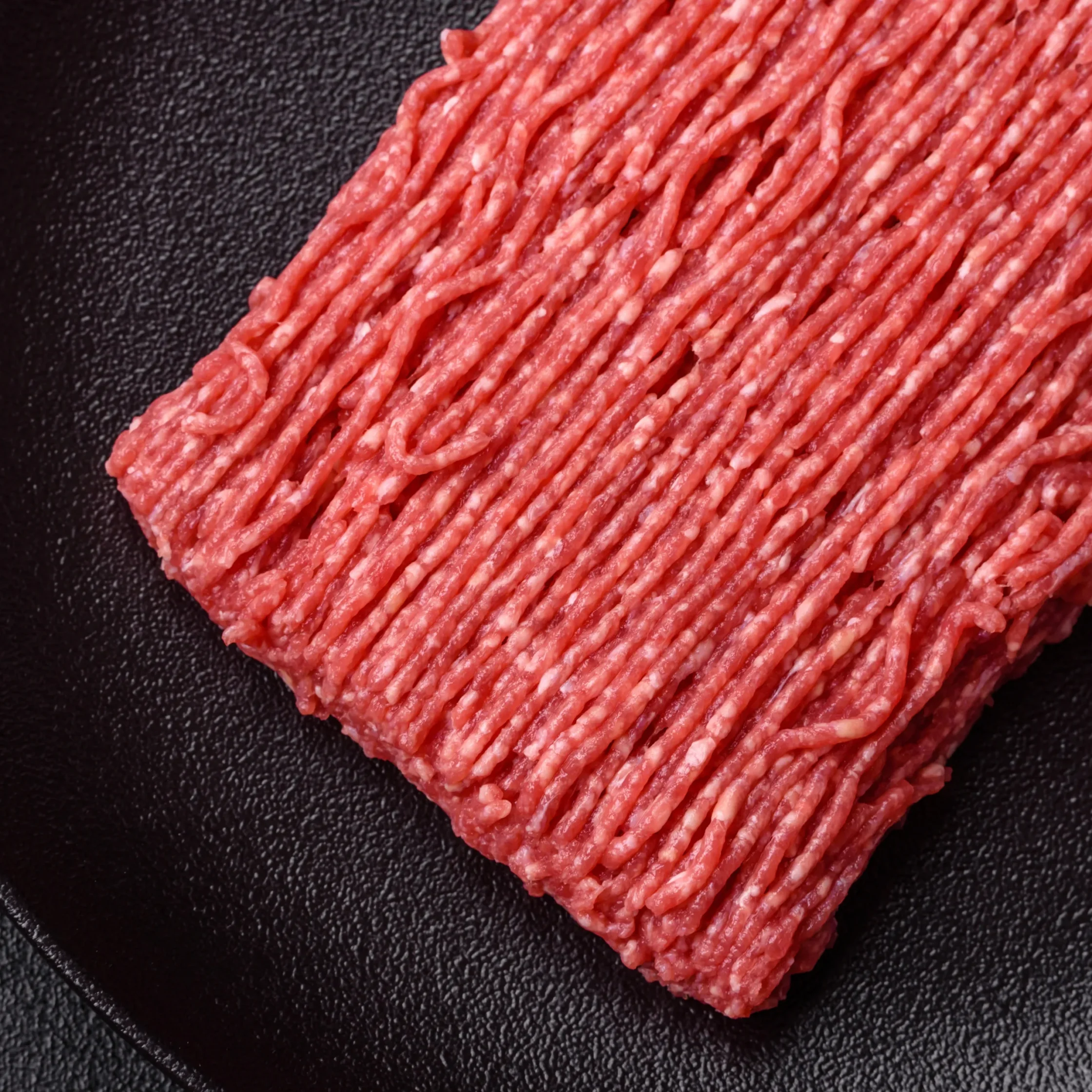 Close-up of a raw ground beef patty with ridged strands and marbled texture, resting on a black surface; bold red color conveys freshness.