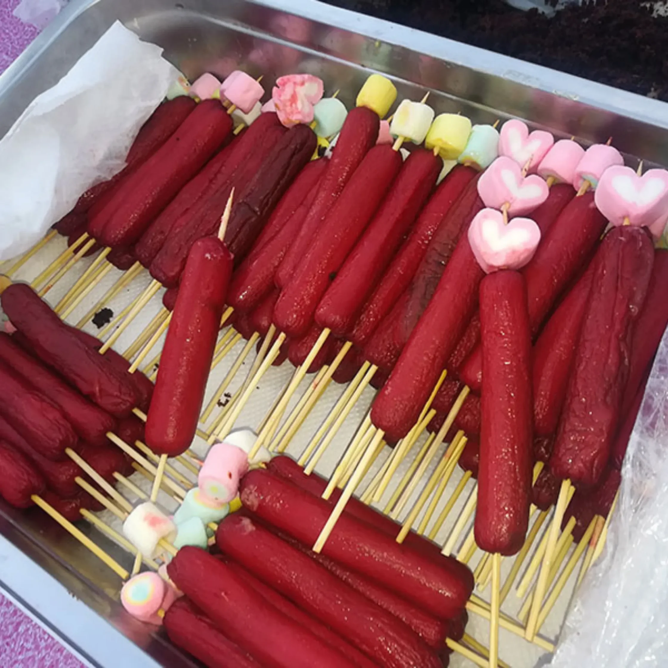 Metal tray filled with red sausage skewers, colorful marshmallows, and pink heart-shaped sweets, creating a playful, festive market display.
