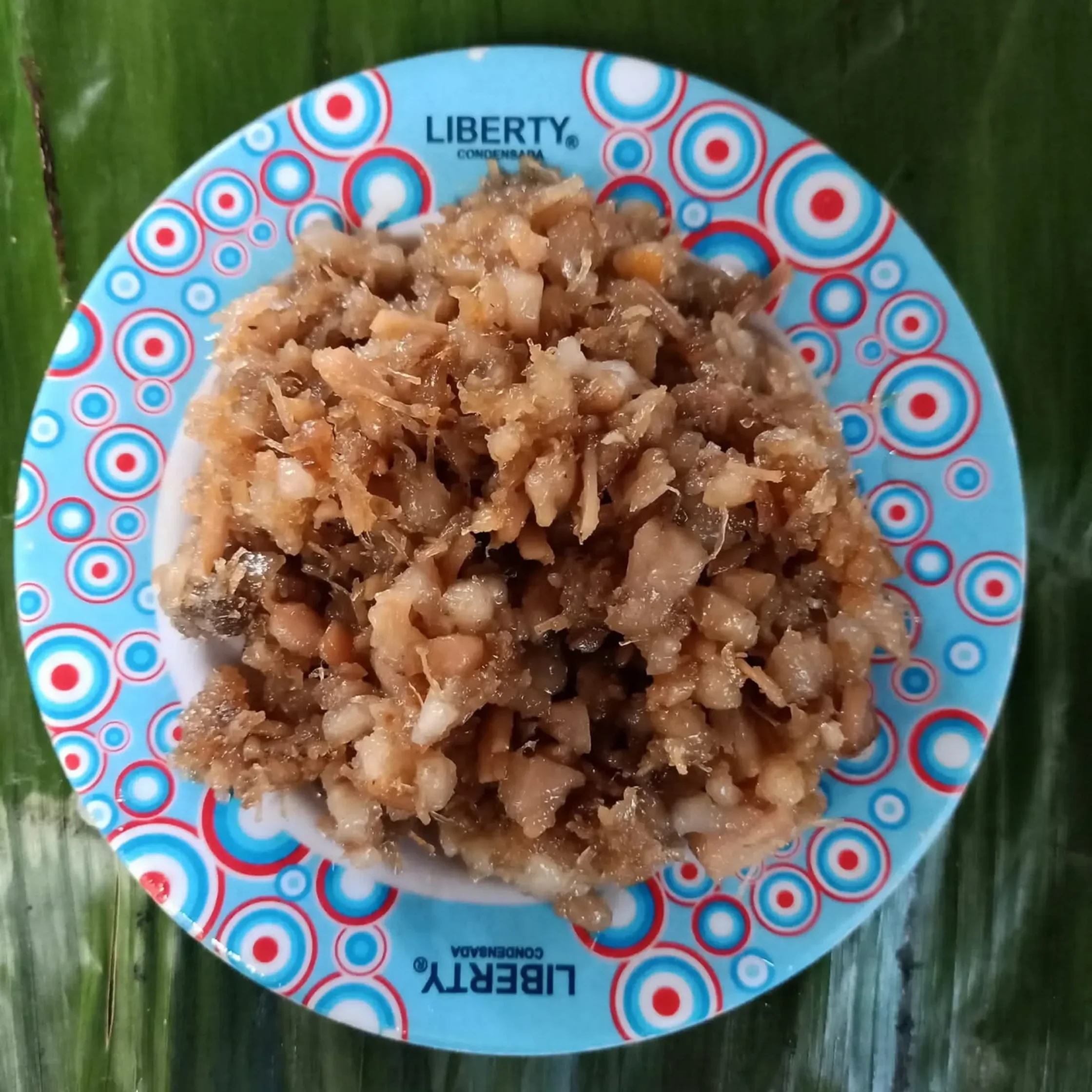 a cooked meat dish on a blue bowl with red circles places on a banana leaf