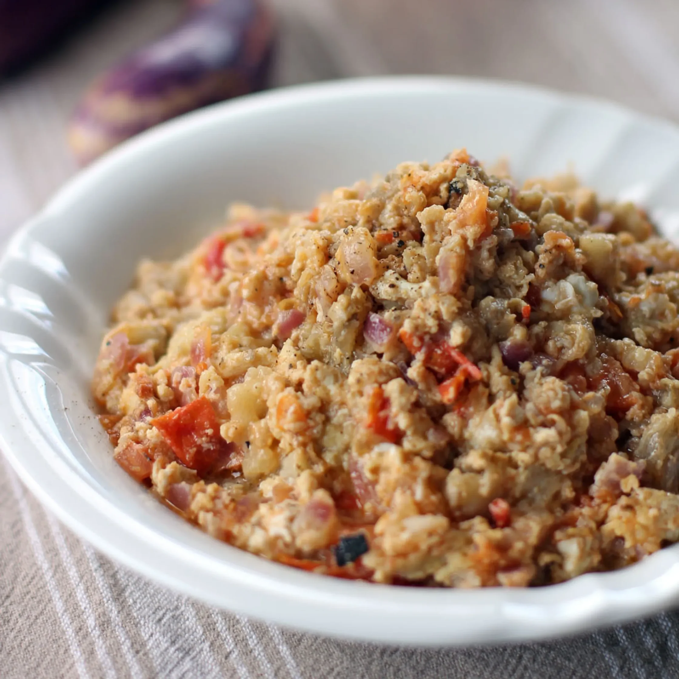 Close-up of a crumbly, baked mixture with visible red peppers in a white bowl on a striped tablecloth.