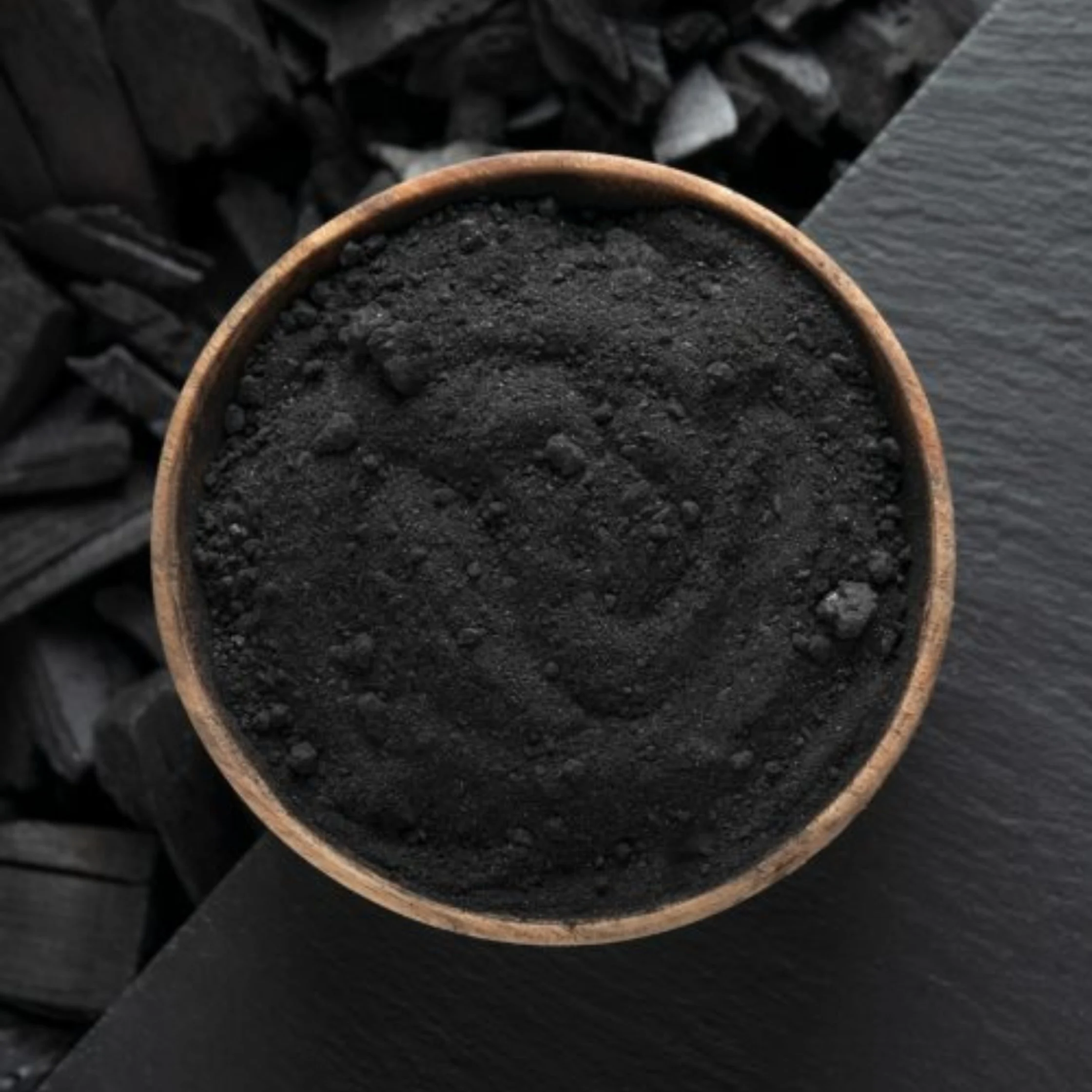 Top-down view of a copper-rimmed bowl filled with coarse black charcoal powder, resting on dark charcoal pieces; the monochrome scene feels stark and earthy.
