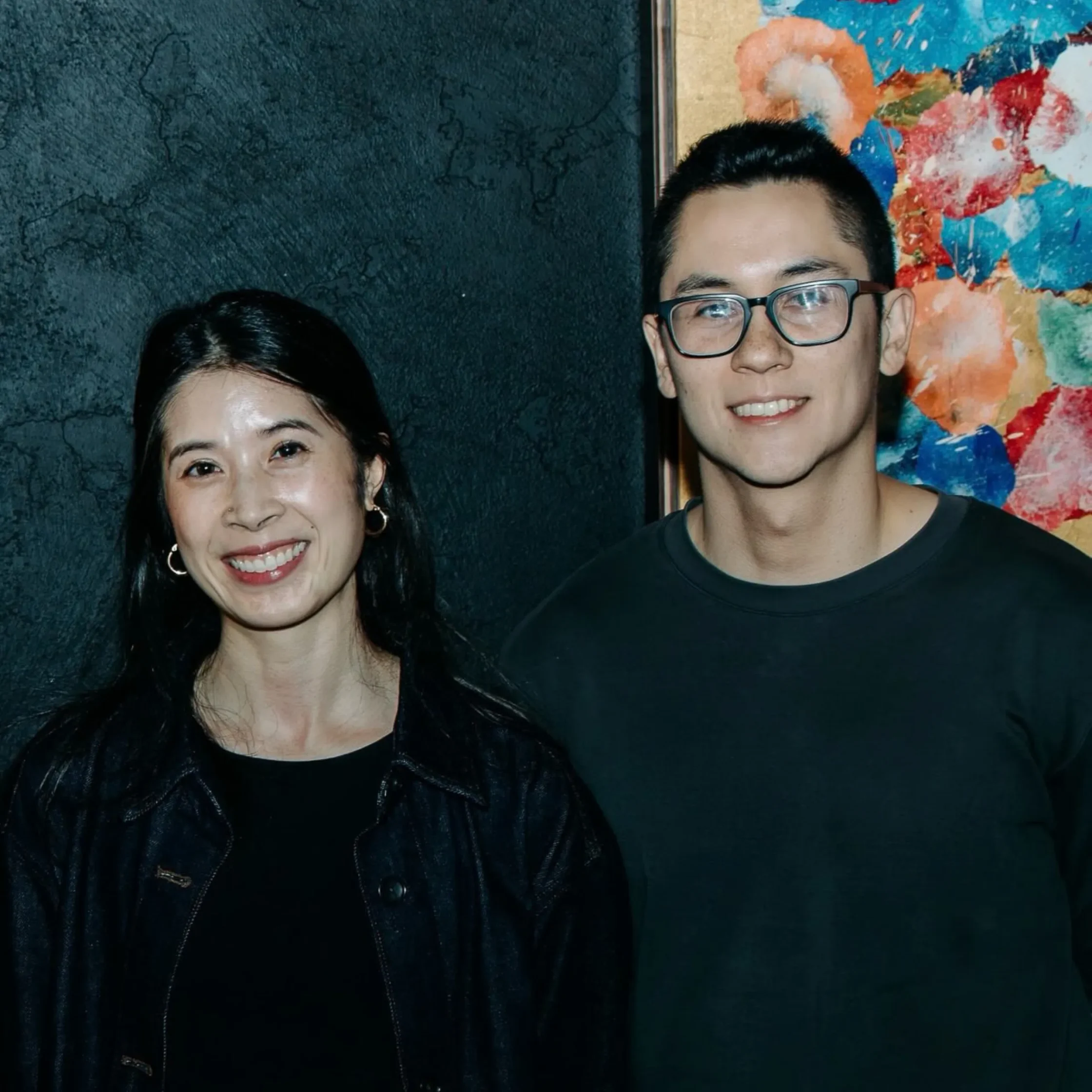 Portrait of a smiling woman and man with glasses standing together against a dark textured wall and colorful abstract painting; friendly mood.