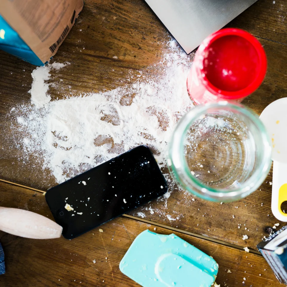 Top-down view of a wooden counter sprinkled with flour, with a red measuring cup, silicone molds, a jar, a bag of flour, and a black smartphone scattered around.