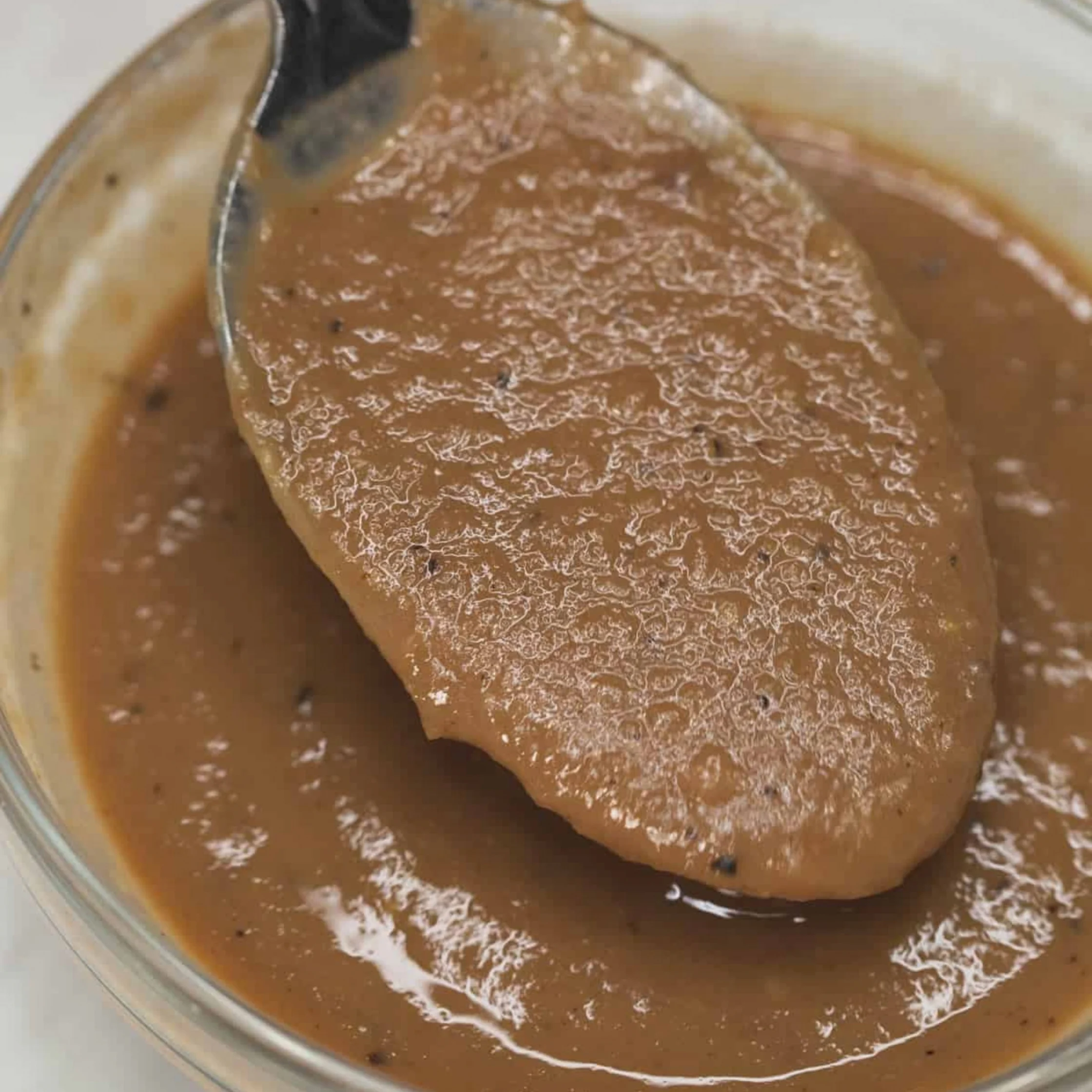 A spoon lifts thick, glossy brown sauce with a slightly grainy texture from a glass bowl; the close-up feels warm and appetizing.