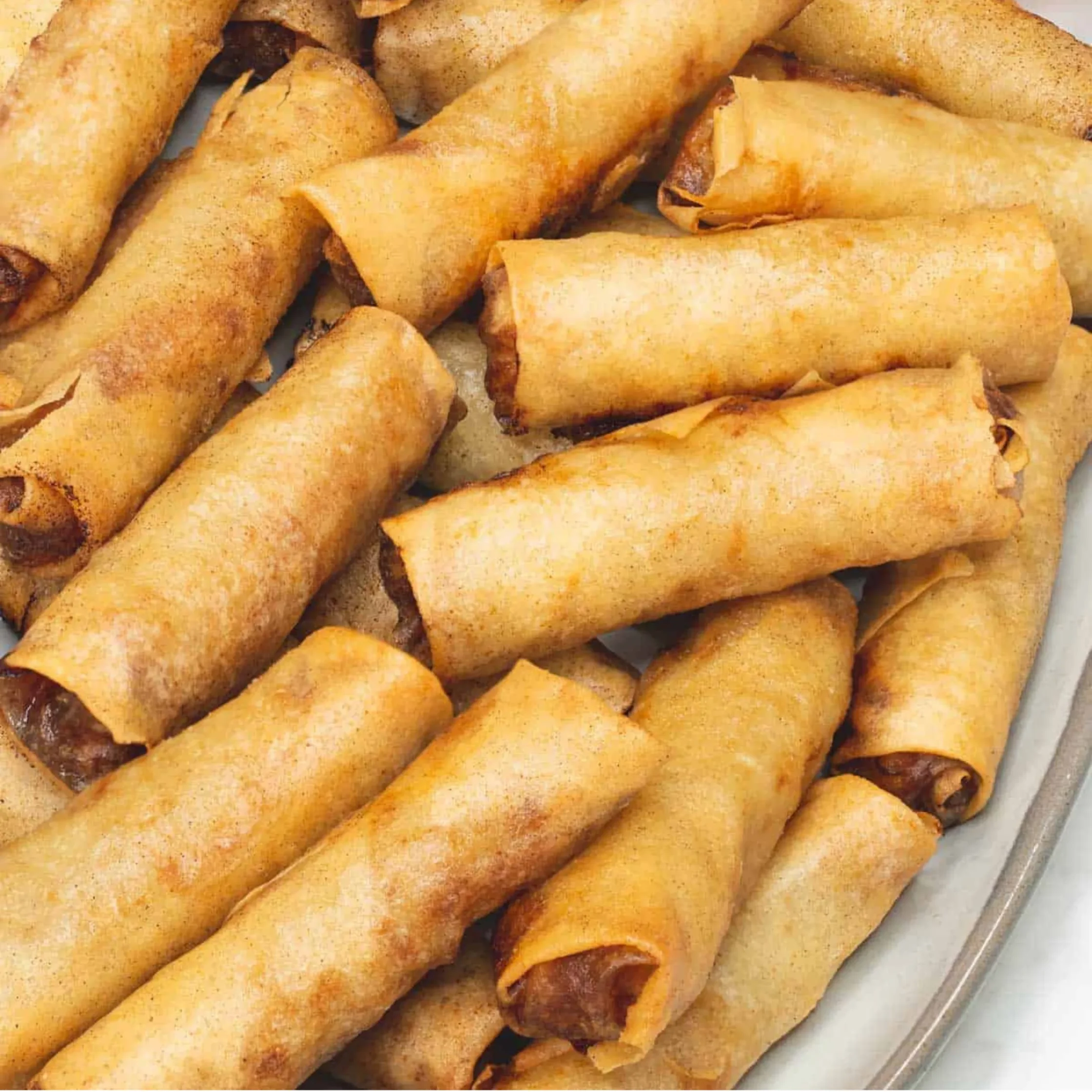 Close-up of golden-brown crispy rolled lumpia or spring rolls piled on a plate, with flaky edges and savory filling visible at the ends.