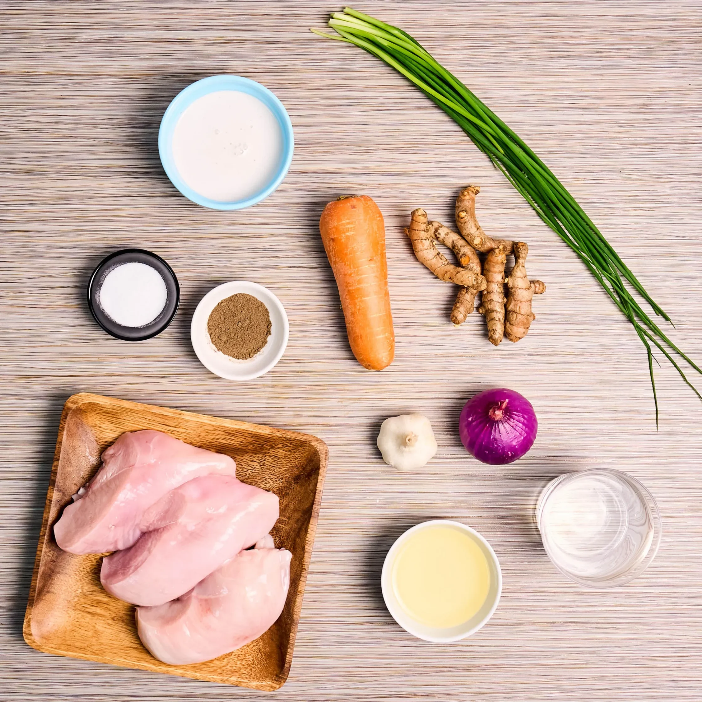 Raw chicken on a wooden cutting board with carrots, ginger, scallions, red onion, garlic, and bowls of salt and spices nearby in a kitchen setting.