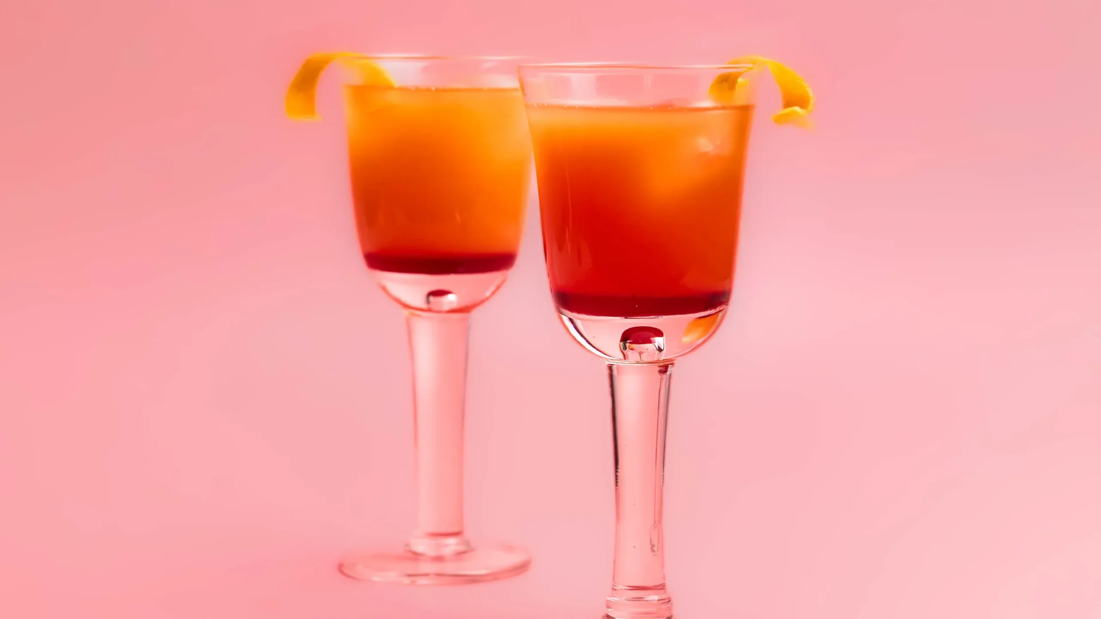 Two champagne flutes filled with orange cocktails and lemon twists, set against a pink backdrop.
