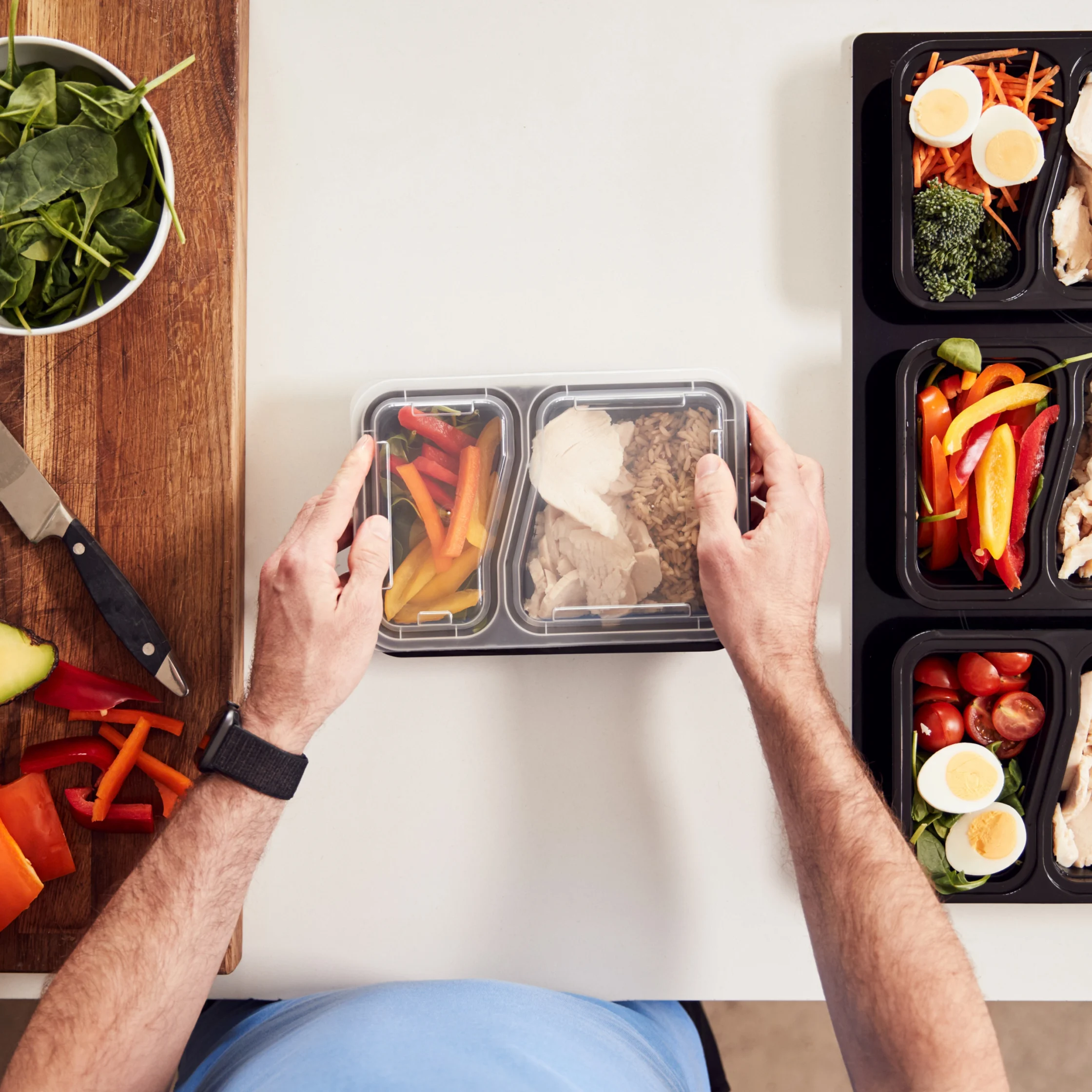 Top-down view of hands packing a meal-prep container with chicken and vegetables, beside colorful prepared meals, salad greens, and cut produce.