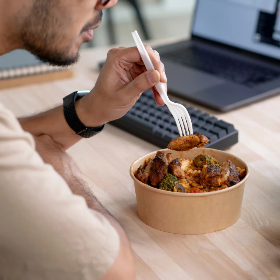 Man uses a white fork to eat a hearty meal from a paper bowl at a desk, with a laptop and keyboard nearby; casual, focused mood.
