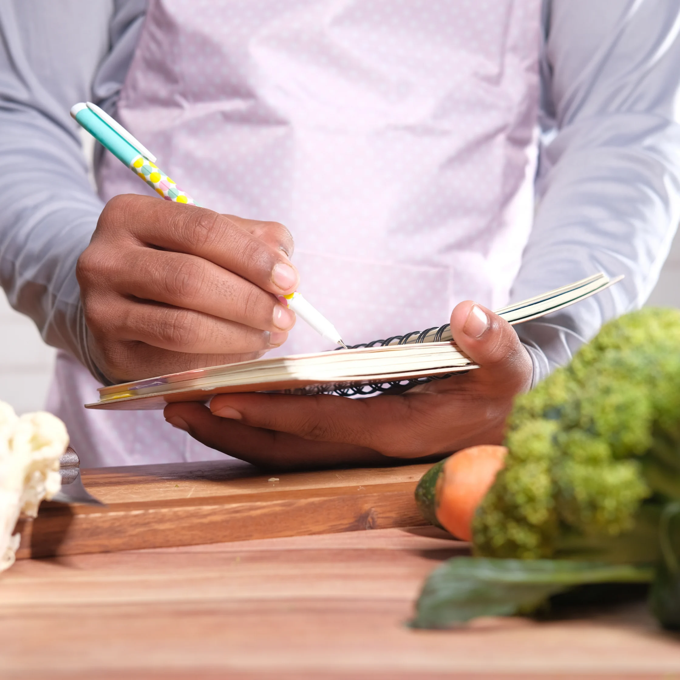 Close-up of a chef writing in a spiral notebook at a wooden counter, surrounded by fresh broccoli, carrots, and greens; focused, hands-on mood.