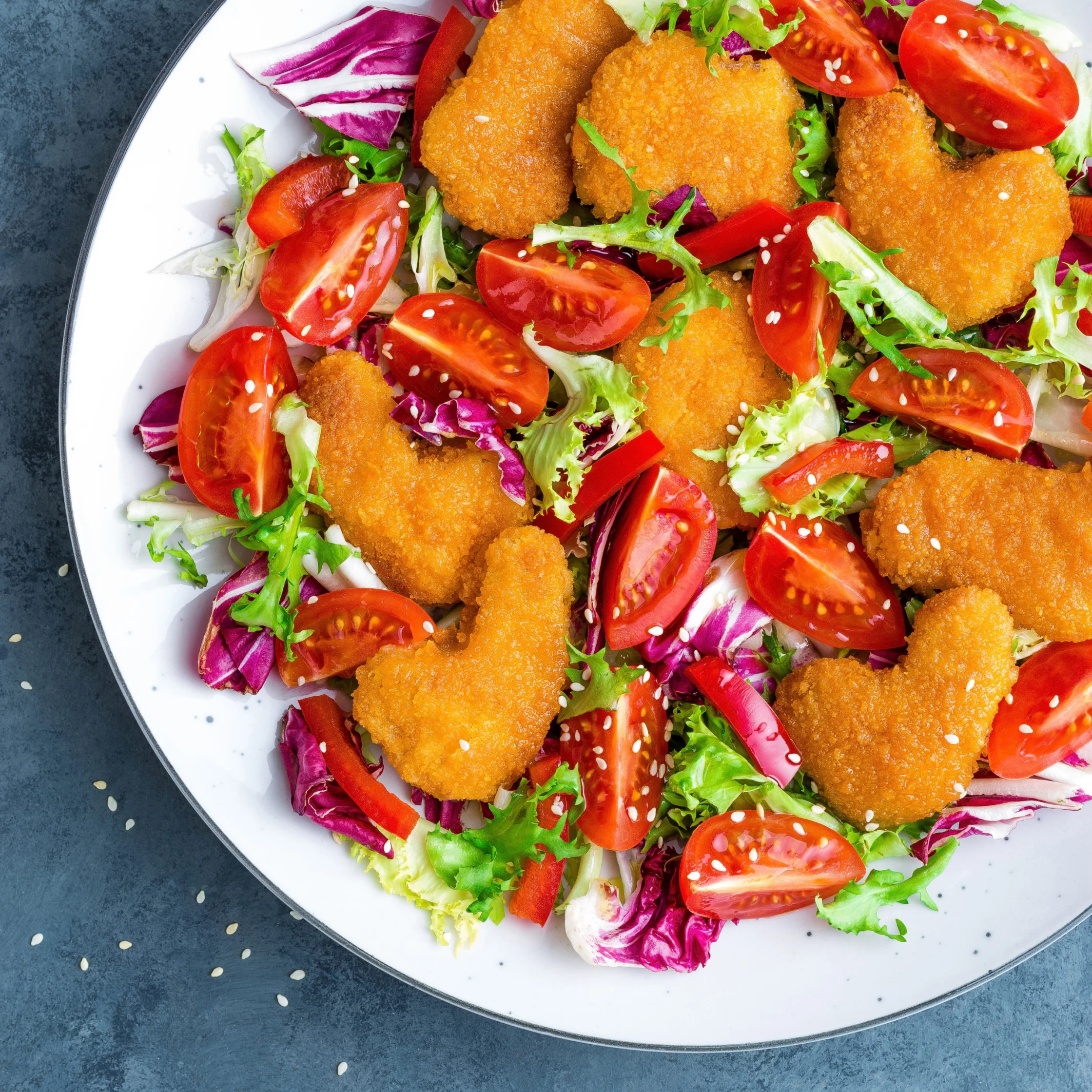 Overhead view of an appetizing salad with golden breaded chicken, cherry tomatoes, leafy greens, red cabbage, peppers, and sesame seeds.