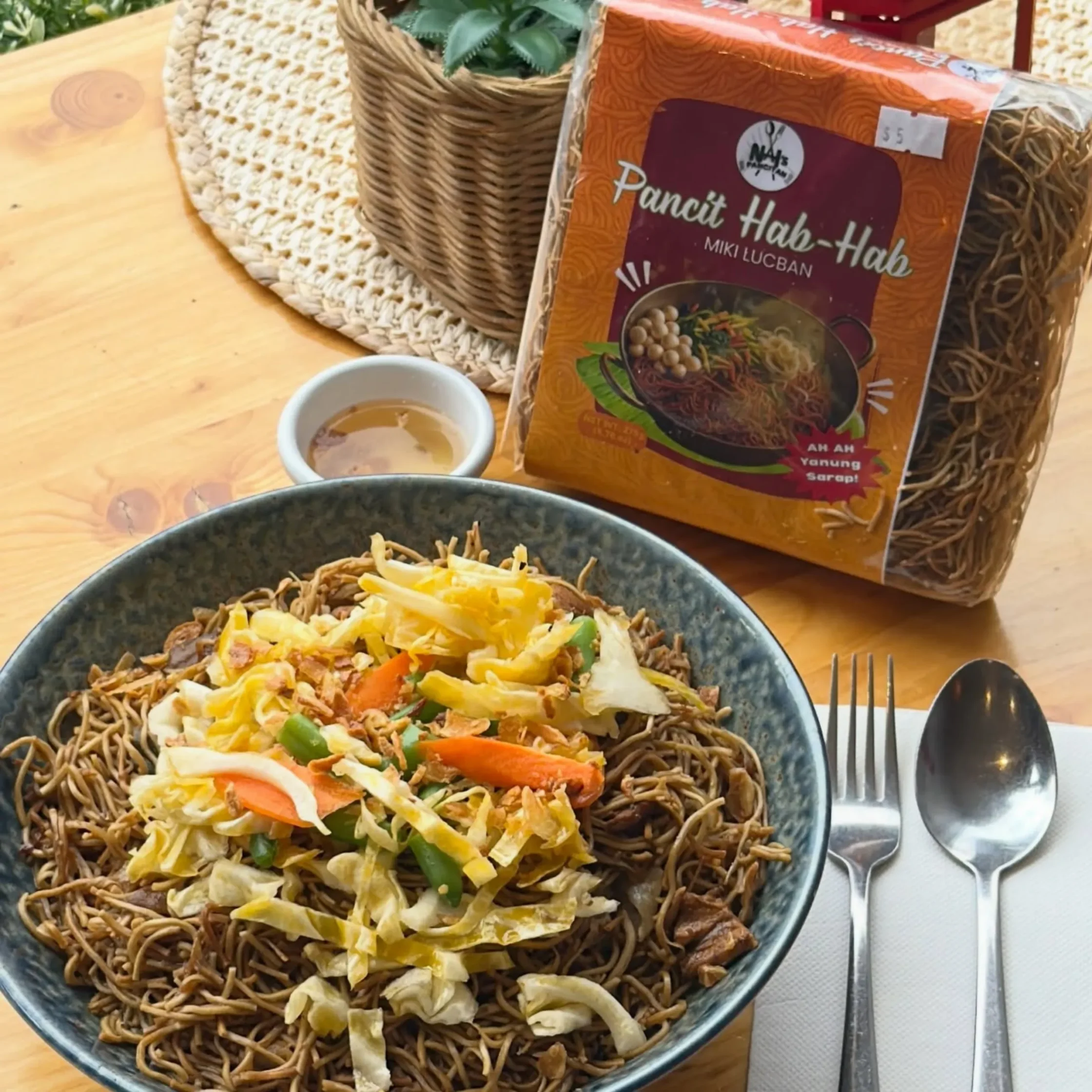 Bowl of fried noodles with cabbage and vegetables on a wooden table, with a bag of Pancit Hab-Hab nearby.
