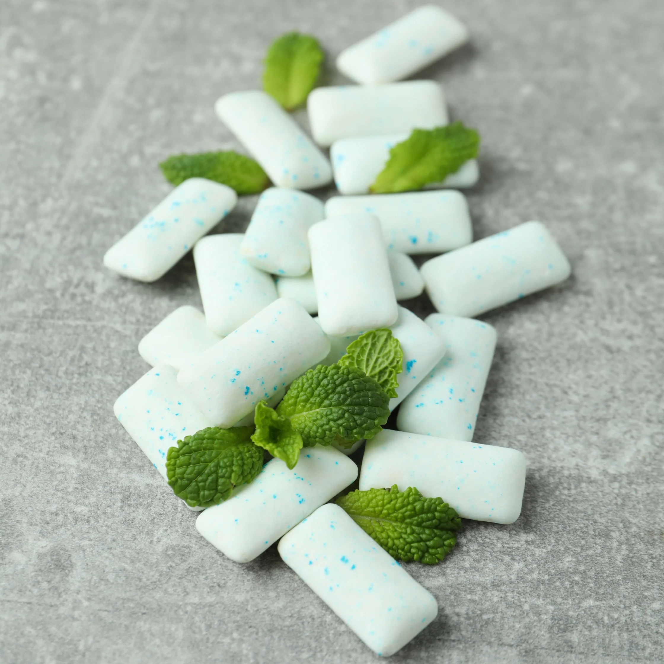 White speckled chewing gum pieces scattered on a gray surface with fresh green mint leaves, creating a cool, refreshing still life.