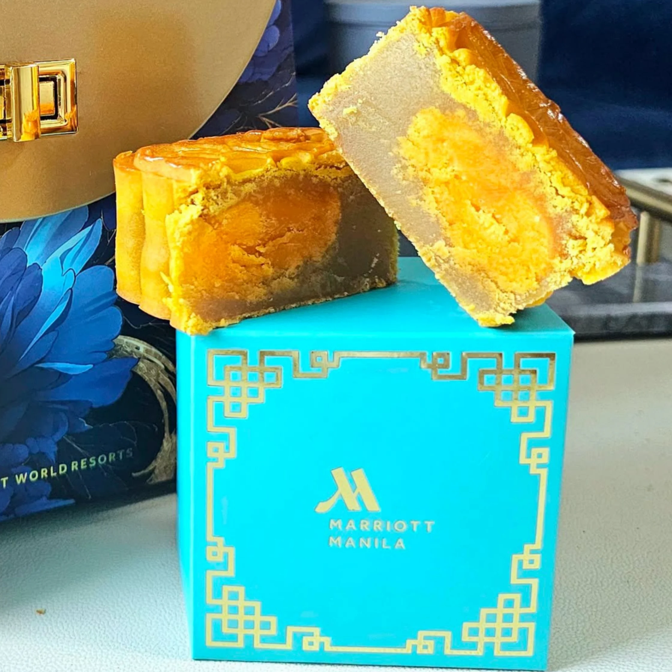 Two golden mooncakes rest on a turquoise Marriott Manila gift box, conveying a festive, luxurious mood with rich pastry textures and colors.