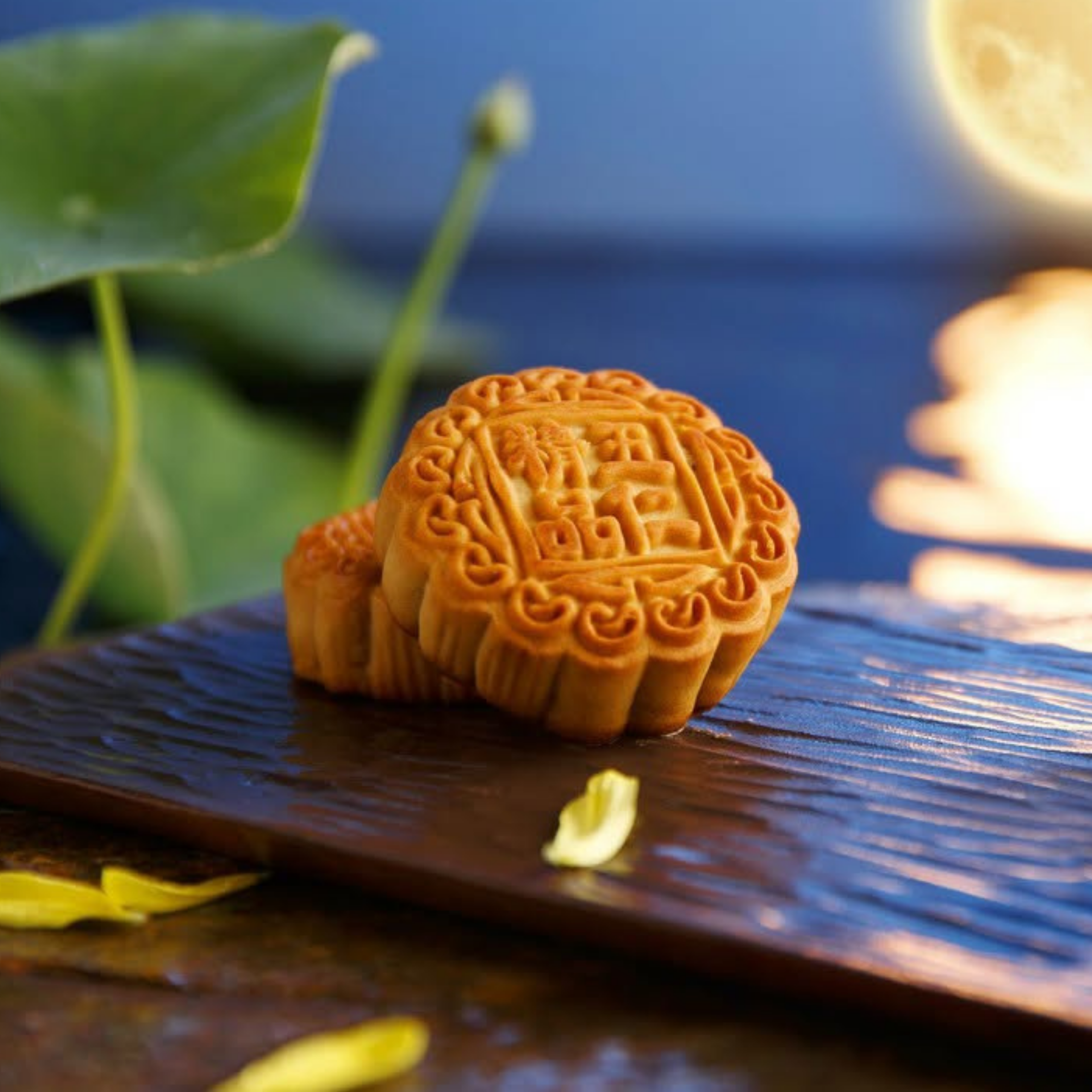 Golden mooncake with ornate floral carving rests on a dark wooden board beside lotus leaves and petals, glowing in warm moonlight over blue water.